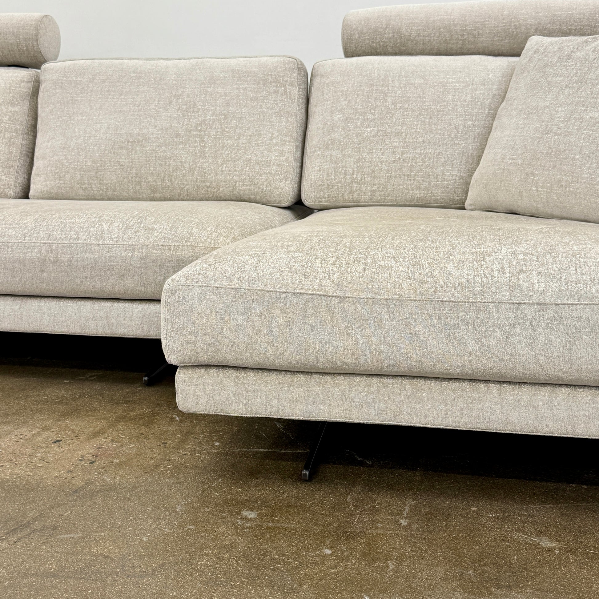 The Poliform Mondrian 2 piece Sectional Sofa with Headrests by Poliform, in light beige, features modern minimalist style with adjustable headrests and a black throw pillow, set on a concrete floor against a plain white wall.