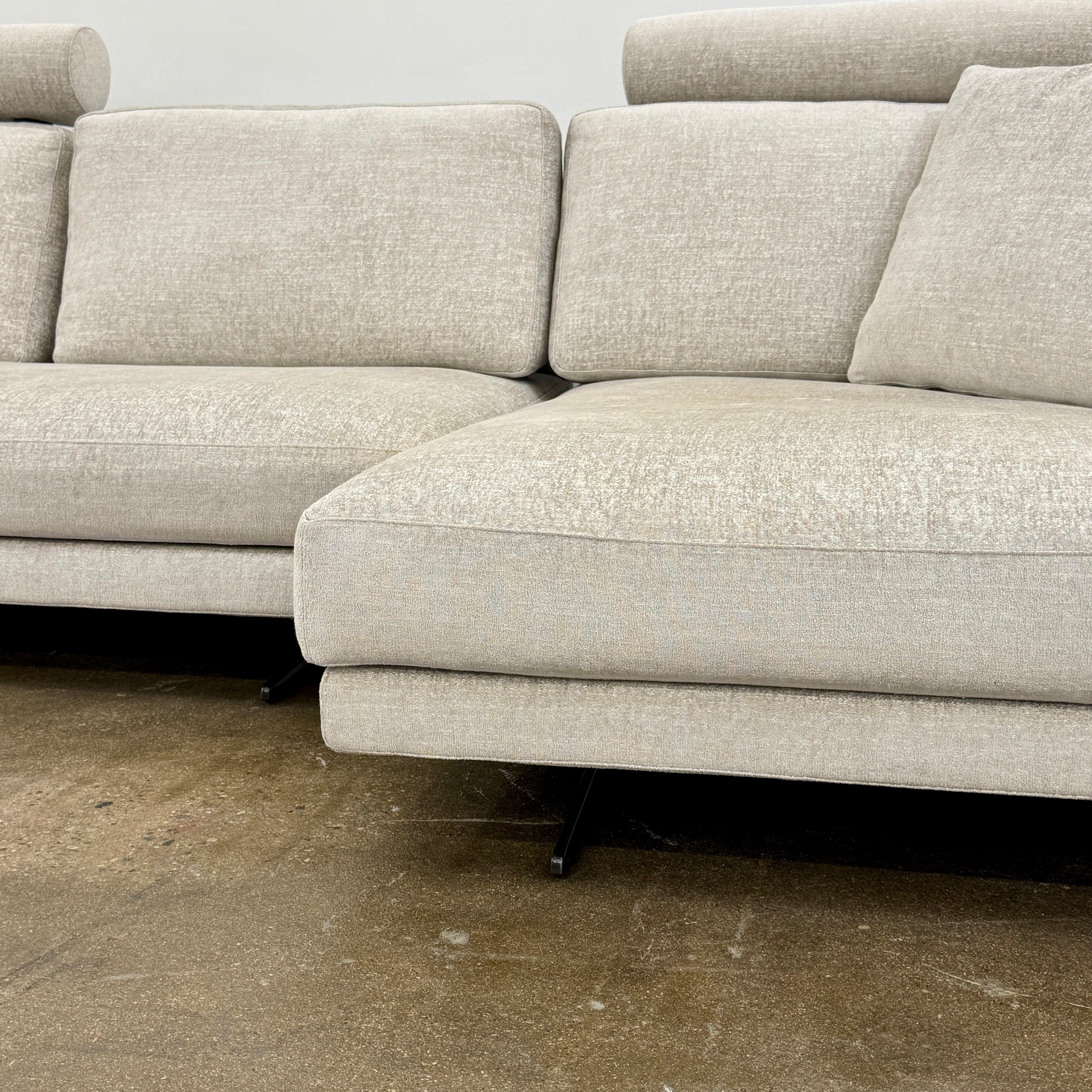 The Poliform Mondrian 2 piece Sectional Sofa with Headrests by Poliform, in light beige, features modern minimalist style with adjustable headrests and a black throw pillow, set on a concrete floor against a plain white wall.