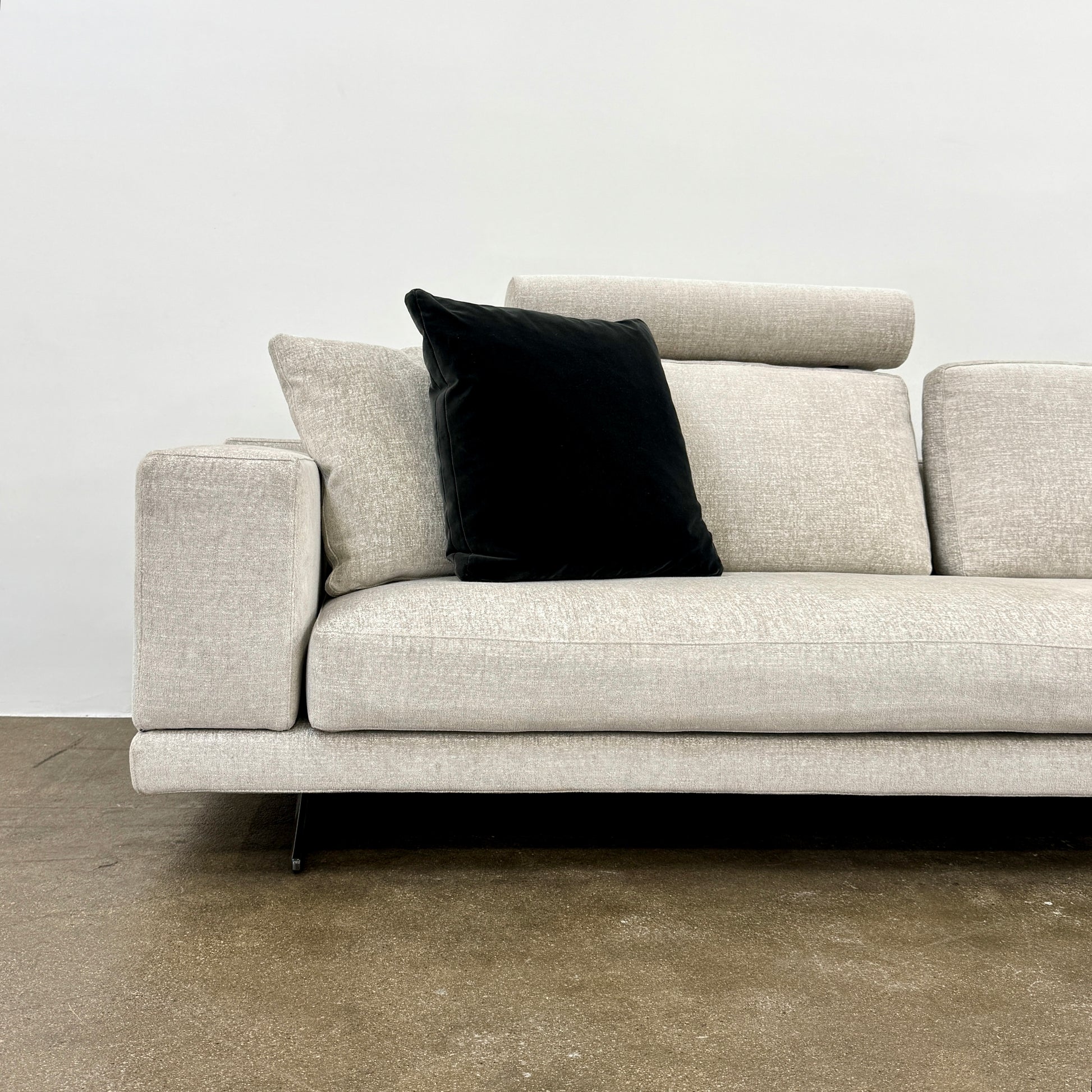 The Poliform Mondrian 2 piece Sectional Sofa with Headrests by Poliform, in light beige, features modern minimalist style with adjustable headrests and a black throw pillow, set on a concrete floor against a plain white wall.