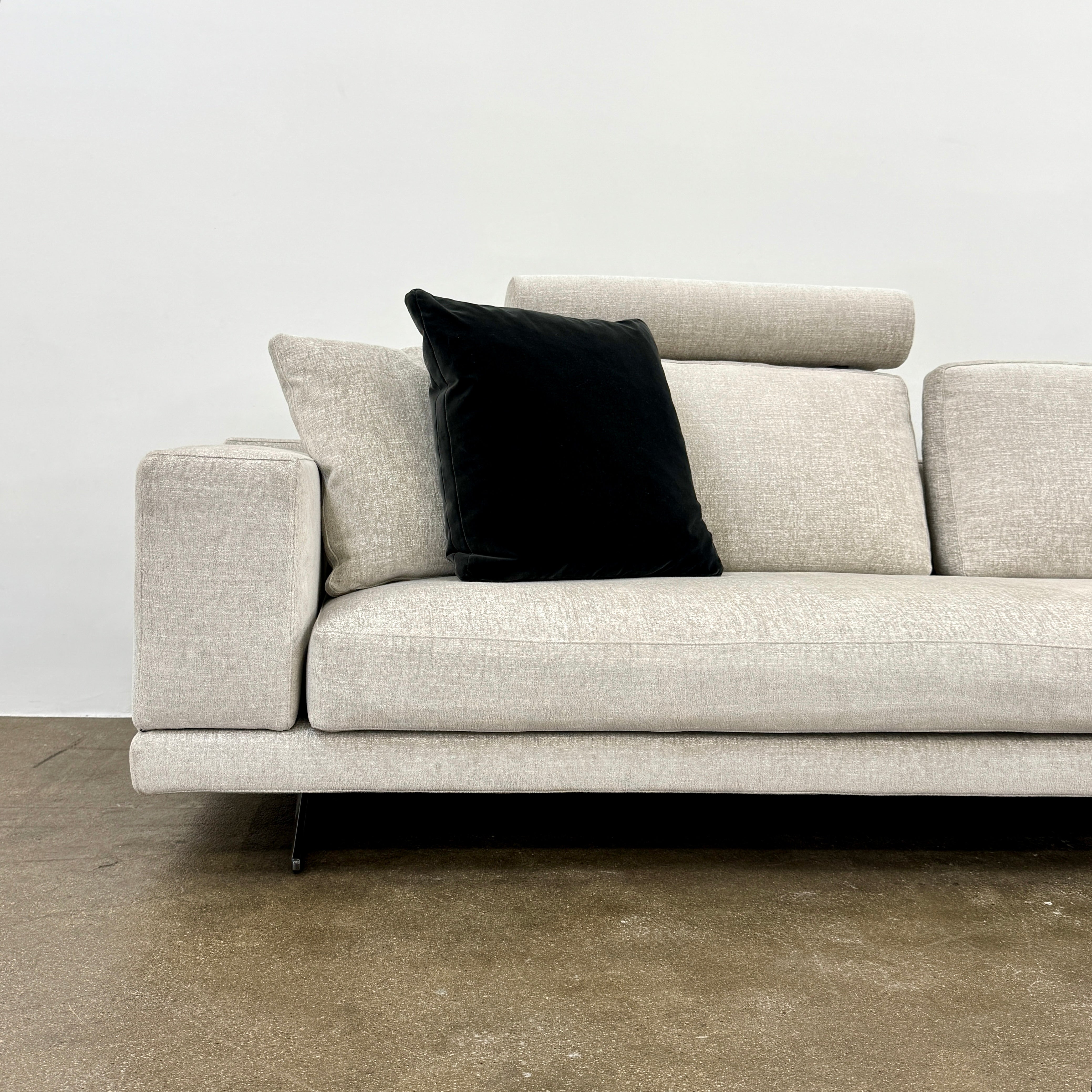 The Poliform Mondrian 2 piece Sectional Sofa with Headrests by Poliform, in light beige, features modern minimalist style with adjustable headrests and a black throw pillow, set on a concrete floor against a plain white wall.