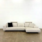 The Poliform Mondrian 2 piece Sectional Sofa with Headrests by Poliform, in light beige, features modern minimalist style with adjustable headrests and a black throw pillow, set on a concrete floor against a plain white wall.