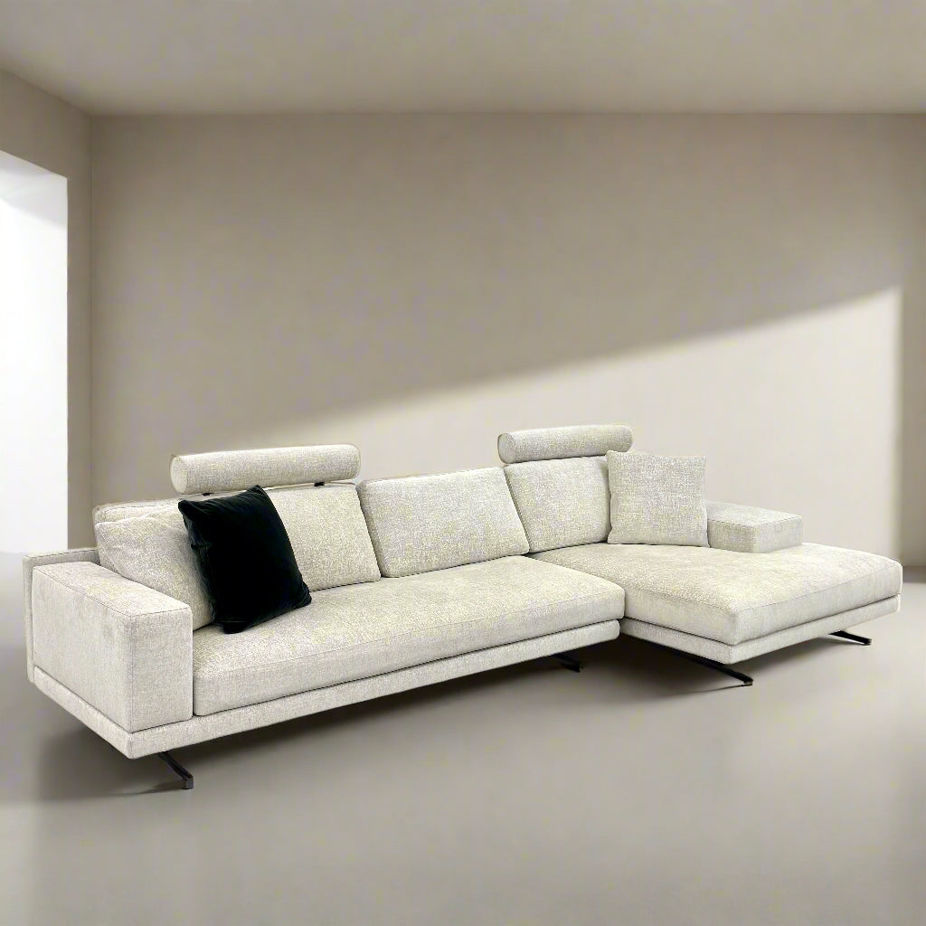 The Poliform Mondrian 2 piece Sectional Sofa with Headrests by Poliform, in light beige, features modern minimalist style with adjustable headrests and a black throw pillow, set on a concrete floor against a plain white wall.