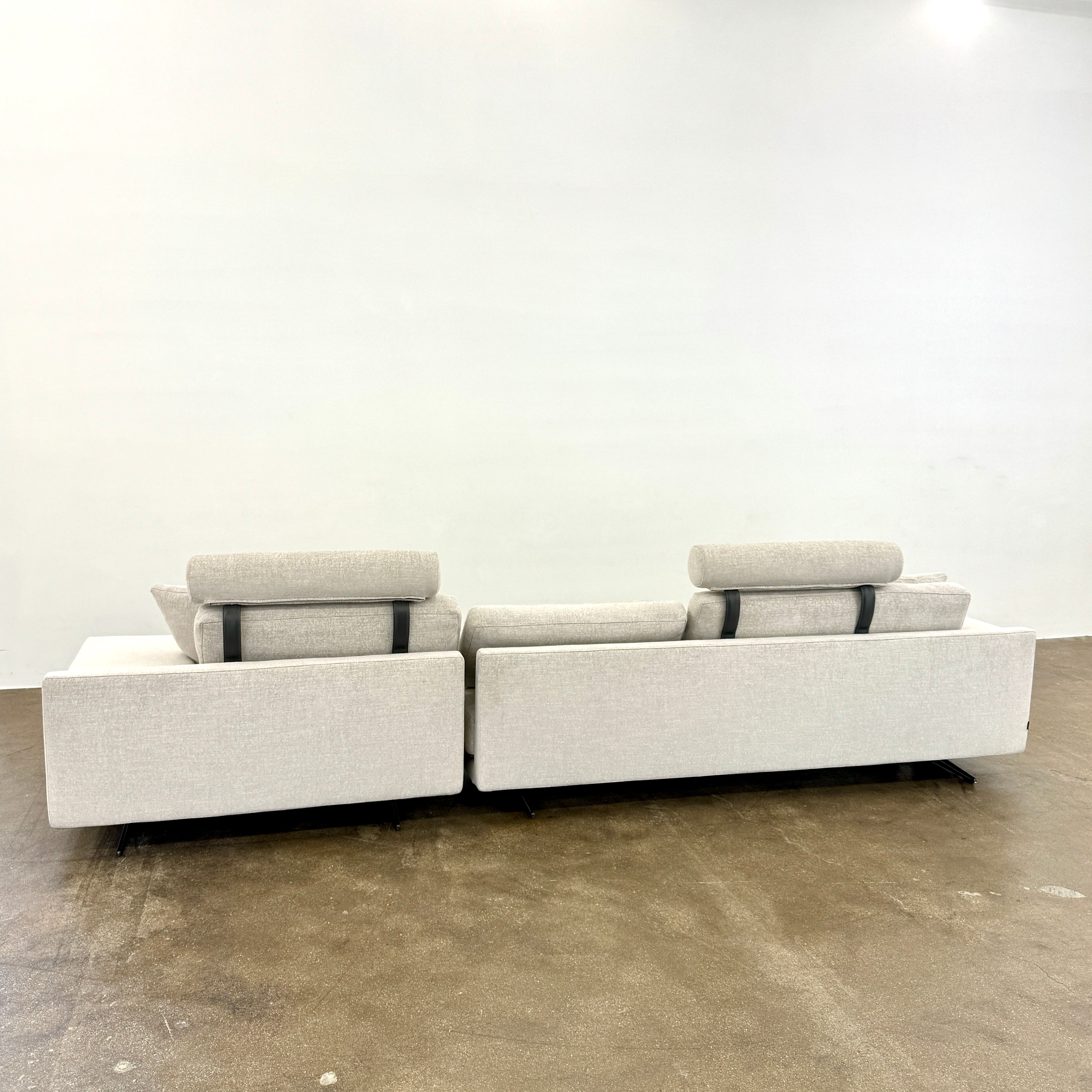 The Poliform Mondrian 2 piece Sectional Sofa with Headrests by Poliform, in light beige, features modern minimalist style with adjustable headrests and a black throw pillow, set on a concrete floor against a plain white wall.