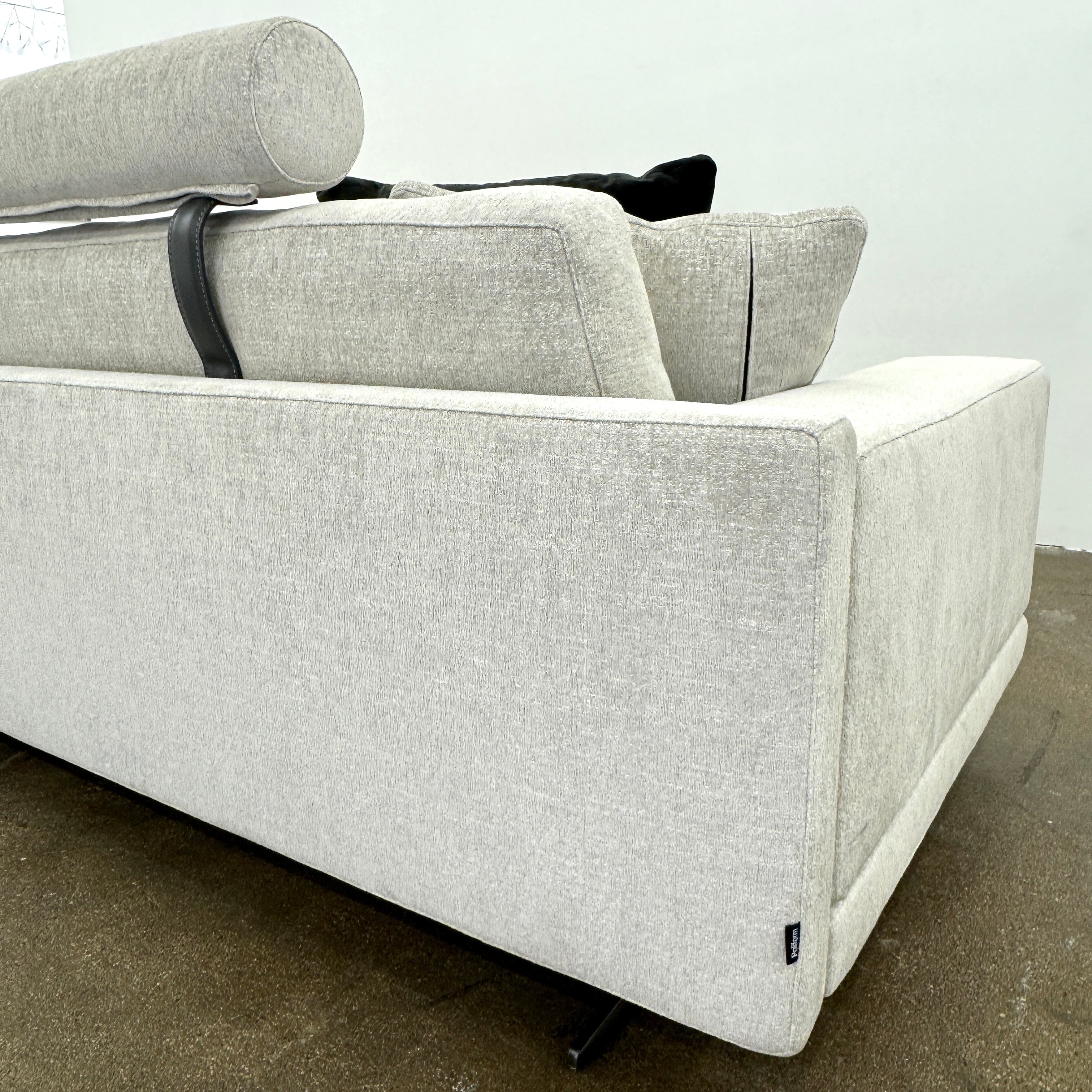 The Poliform Mondrian 2 piece Sectional Sofa with Headrests by Poliform, in light beige, features modern minimalist style with adjustable headrests and a black throw pillow, set on a concrete floor against a plain white wall.