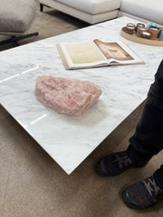 A large Rose Quartz from Modern Resale sits on a white marble table beside an open book and a round tray with jars. A person in black shoes appears partially on the right.