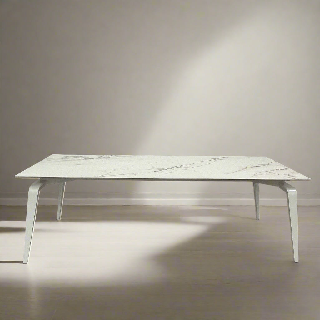 The Ligne Roset Odessa Dining Table showcases a rectangular marble top with angled, folded steel legs, embodying natural minimalism. It is elegantly viewed from the side against a plain background.