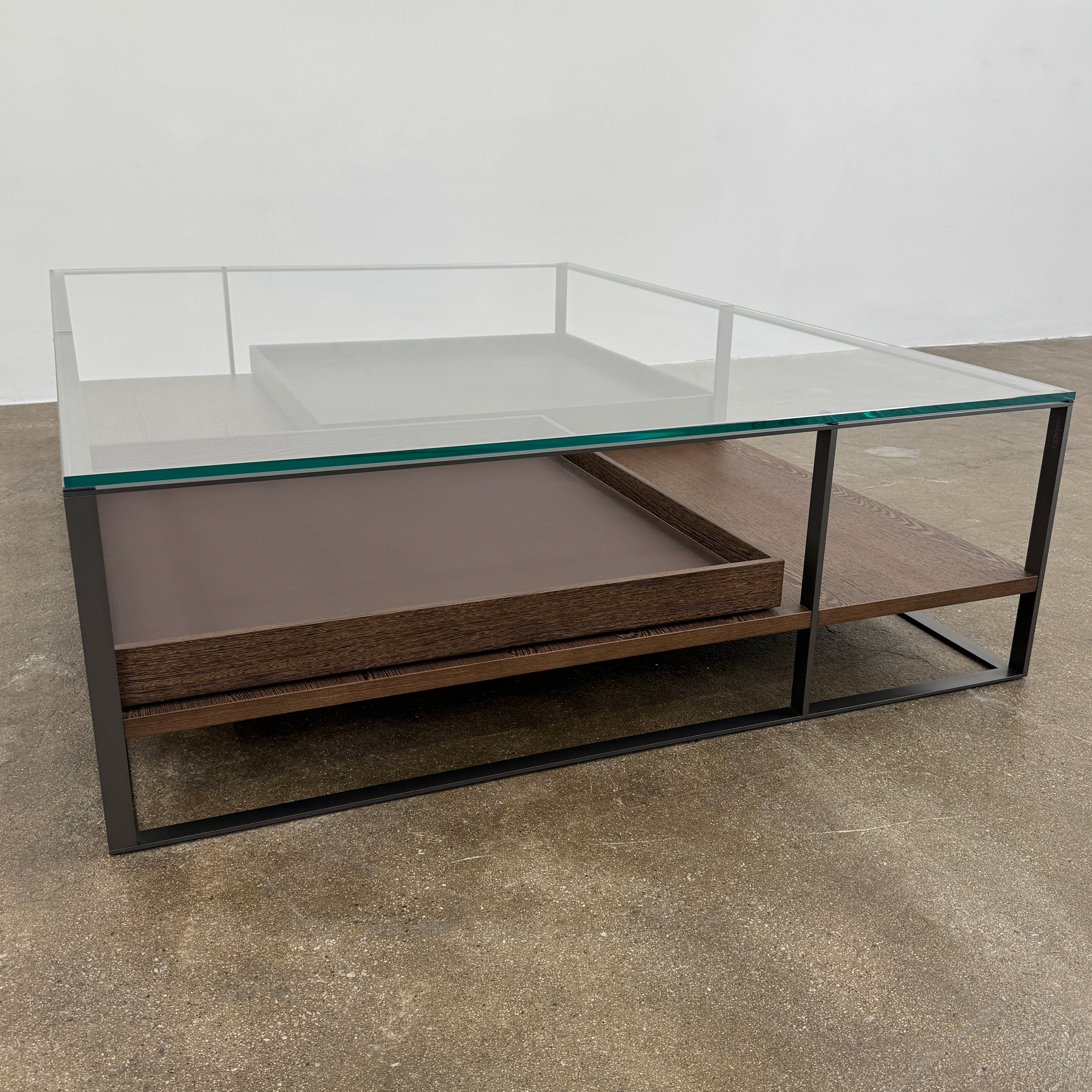The Maxalto Lithos Coffee Table by Maxalto features a square metal frame, glass top, and two wooden lower shelves—one slightly raised—inspired by the original design. Shown against a plain white background.