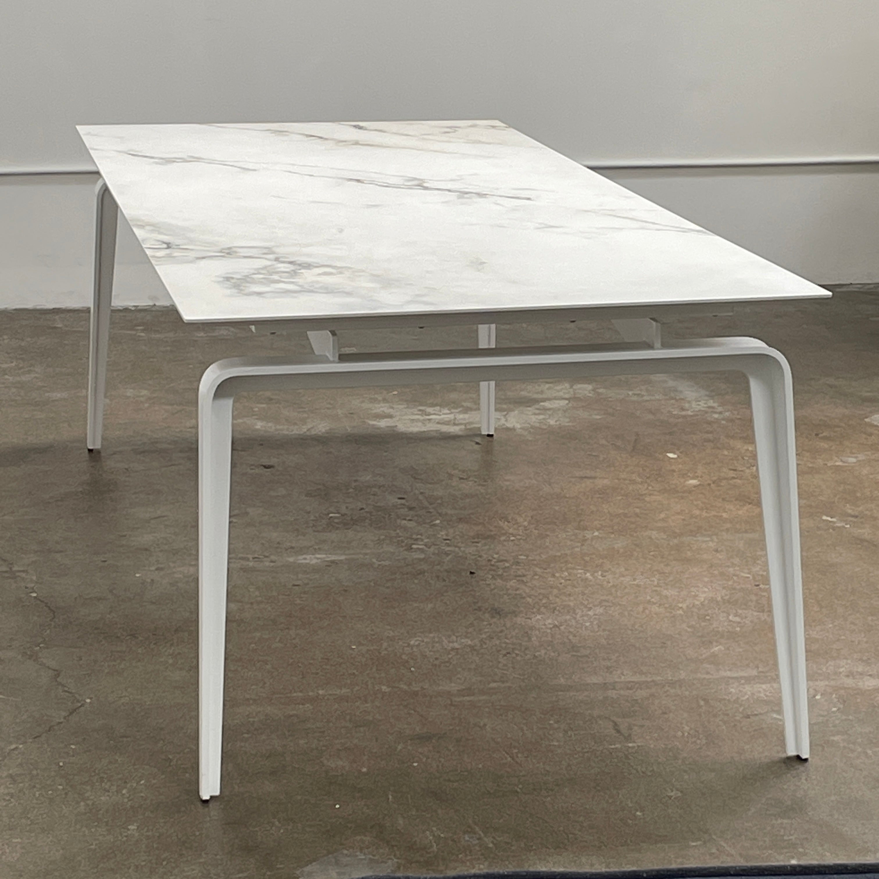 The Ligne Roset Odessa Dining Table showcases a rectangular marble top with angled, folded steel legs, embodying natural minimalism. It is elegantly viewed from the side against a plain background.