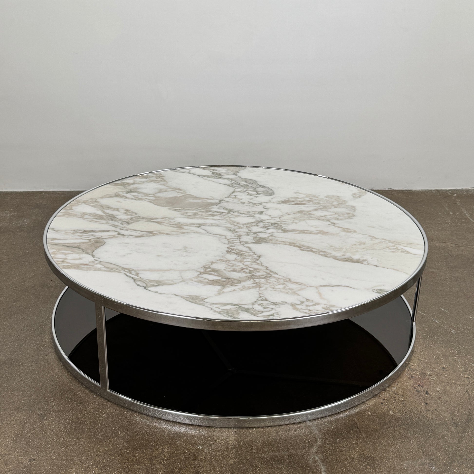 The Minotti Huber Coffee Table by Minotti features a round design with a white statuary marble top, dark lower shelf, and sleek metal frame, set against a plain light background.