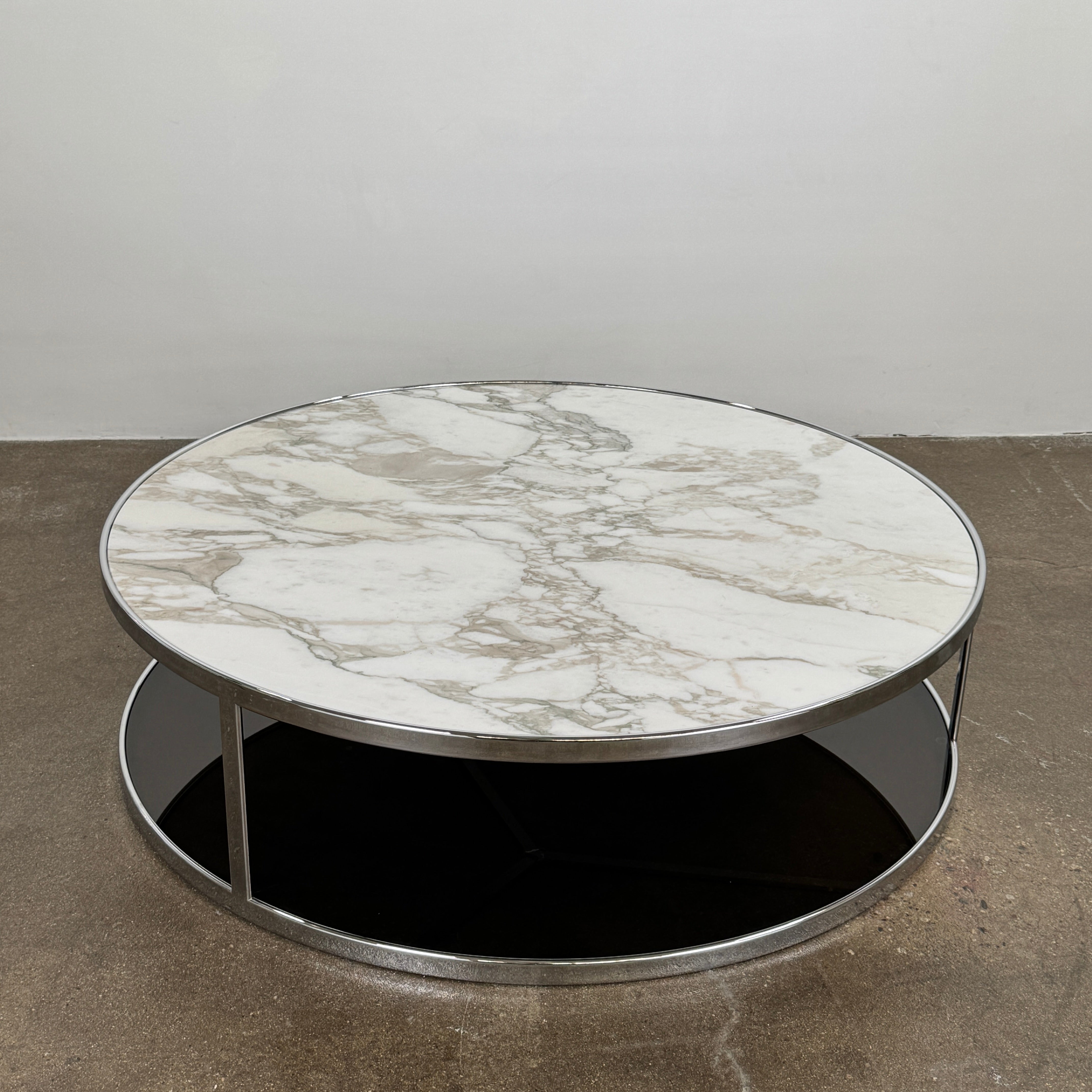 The Minotti Huber Coffee Table by Minotti features a round design with a white statuary marble top, dark lower shelf, and sleek metal frame, set against a plain light background.