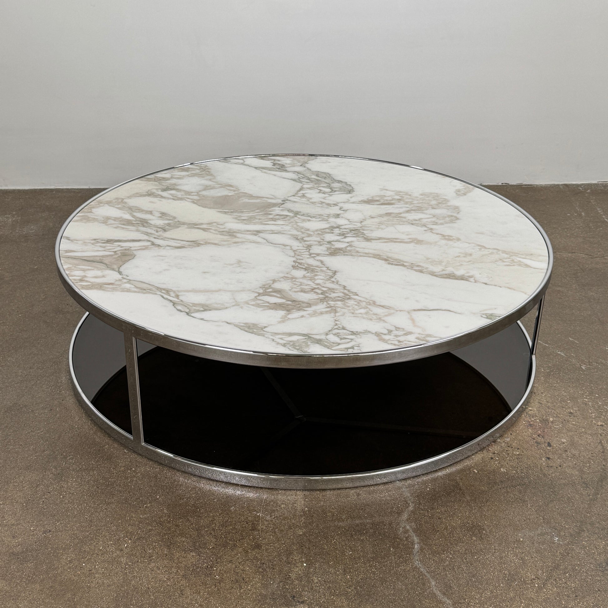 The Minotti Huber Coffee Table by Minotti features a round design with a white statuary marble top, dark lower shelf, and sleek metal frame, set against a plain light background.