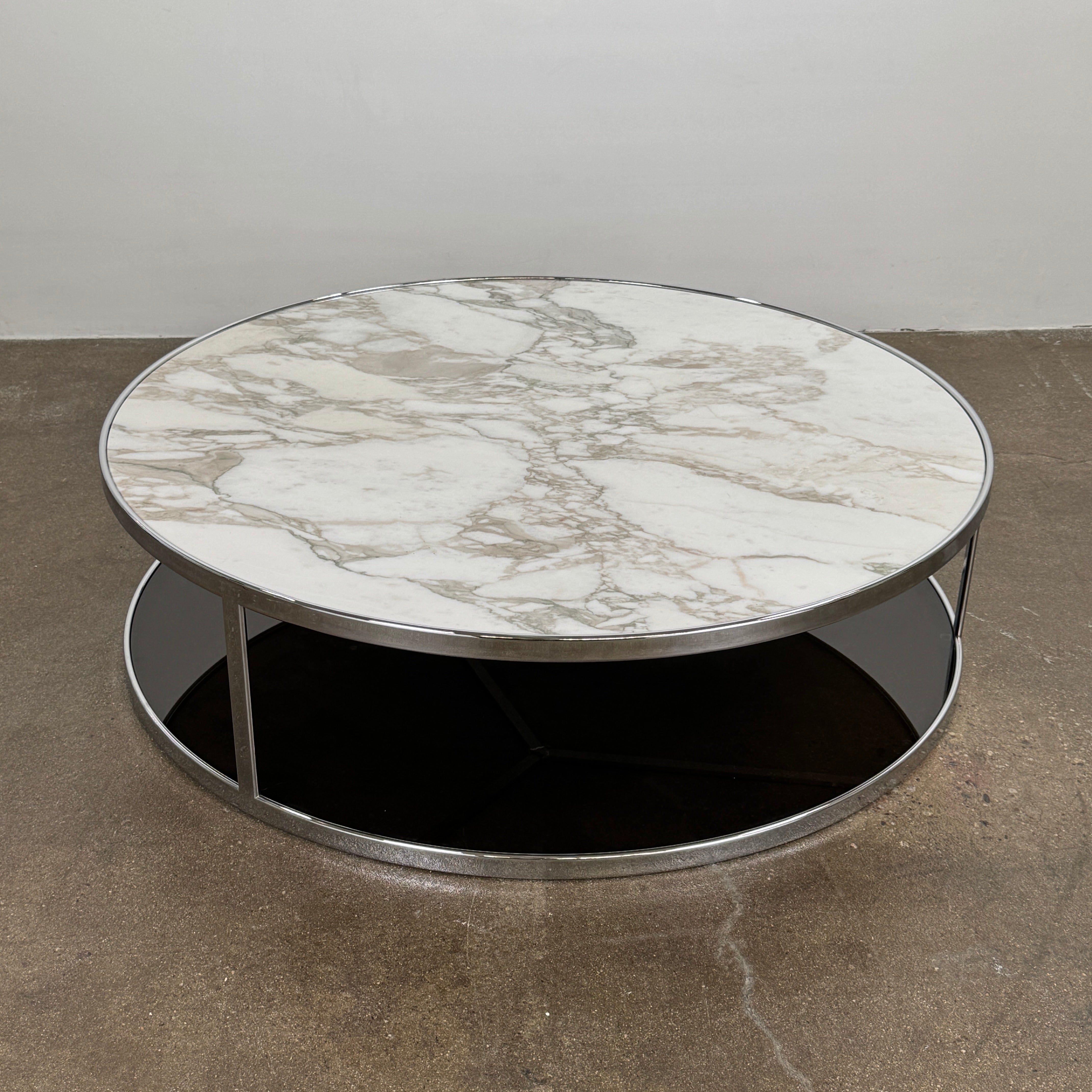 The Minotti Huber Coffee Table by Minotti features a round design with a white statuary marble top, dark lower shelf, and sleek metal frame, set against a plain light background.