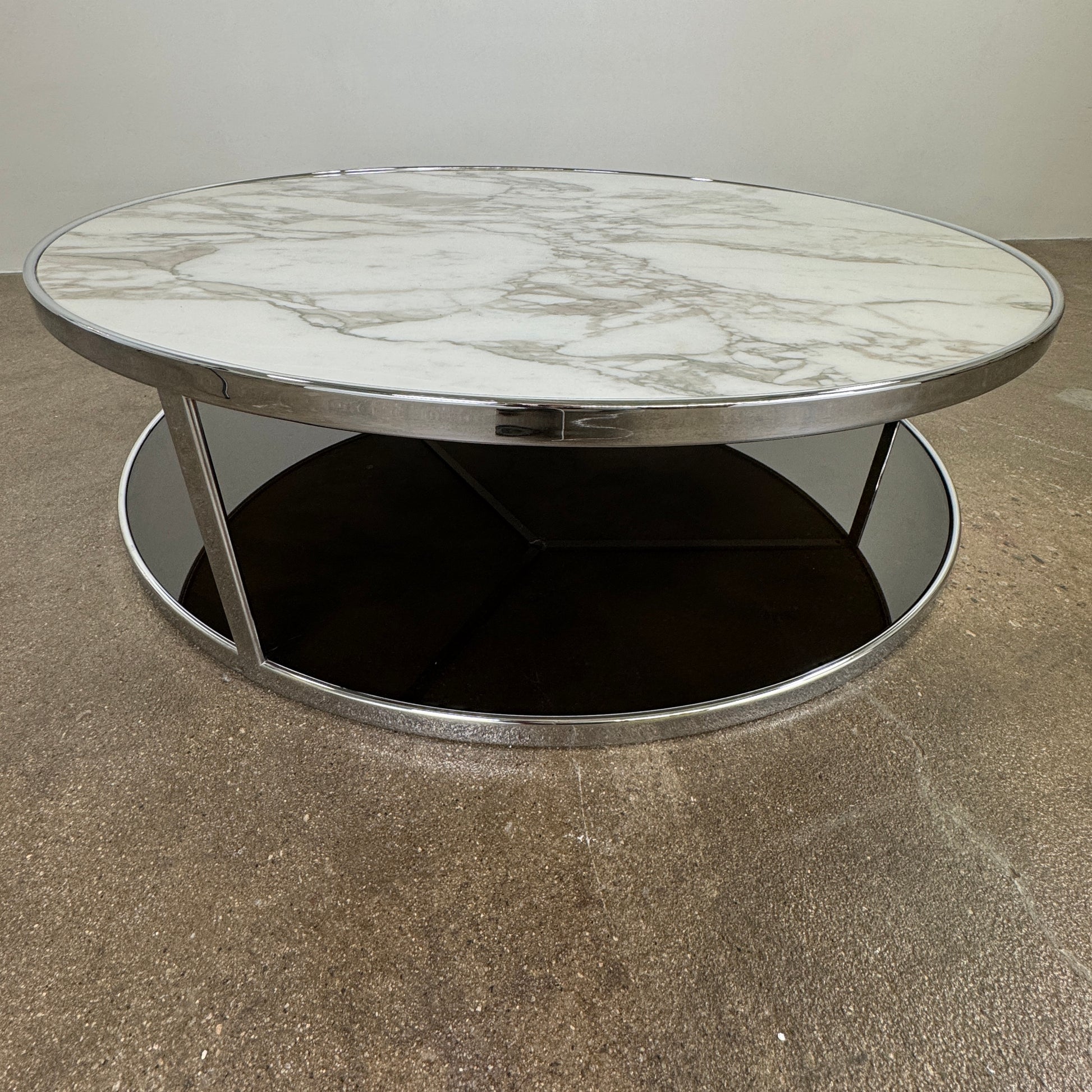 The Minotti Huber Coffee Table by Minotti features a round design with a white statuary marble top, dark lower shelf, and sleek metal frame, set against a plain light background.