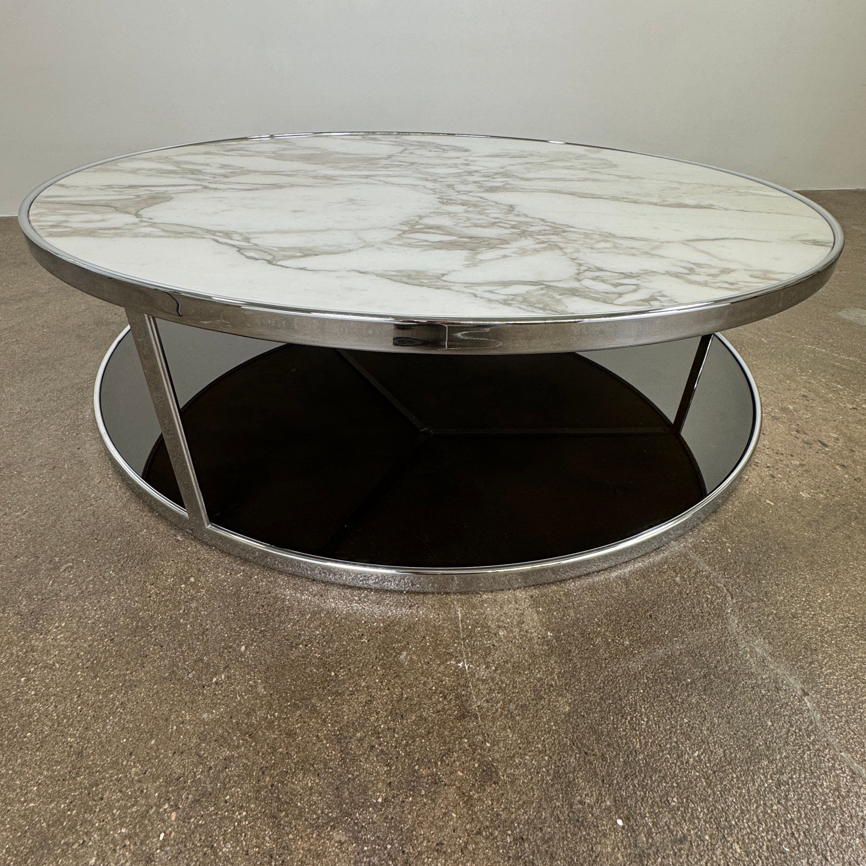The Minotti Huber Coffee Table by Minotti features a round design with a white statuary marble top, dark lower shelf, and sleek metal frame, set against a plain light background.