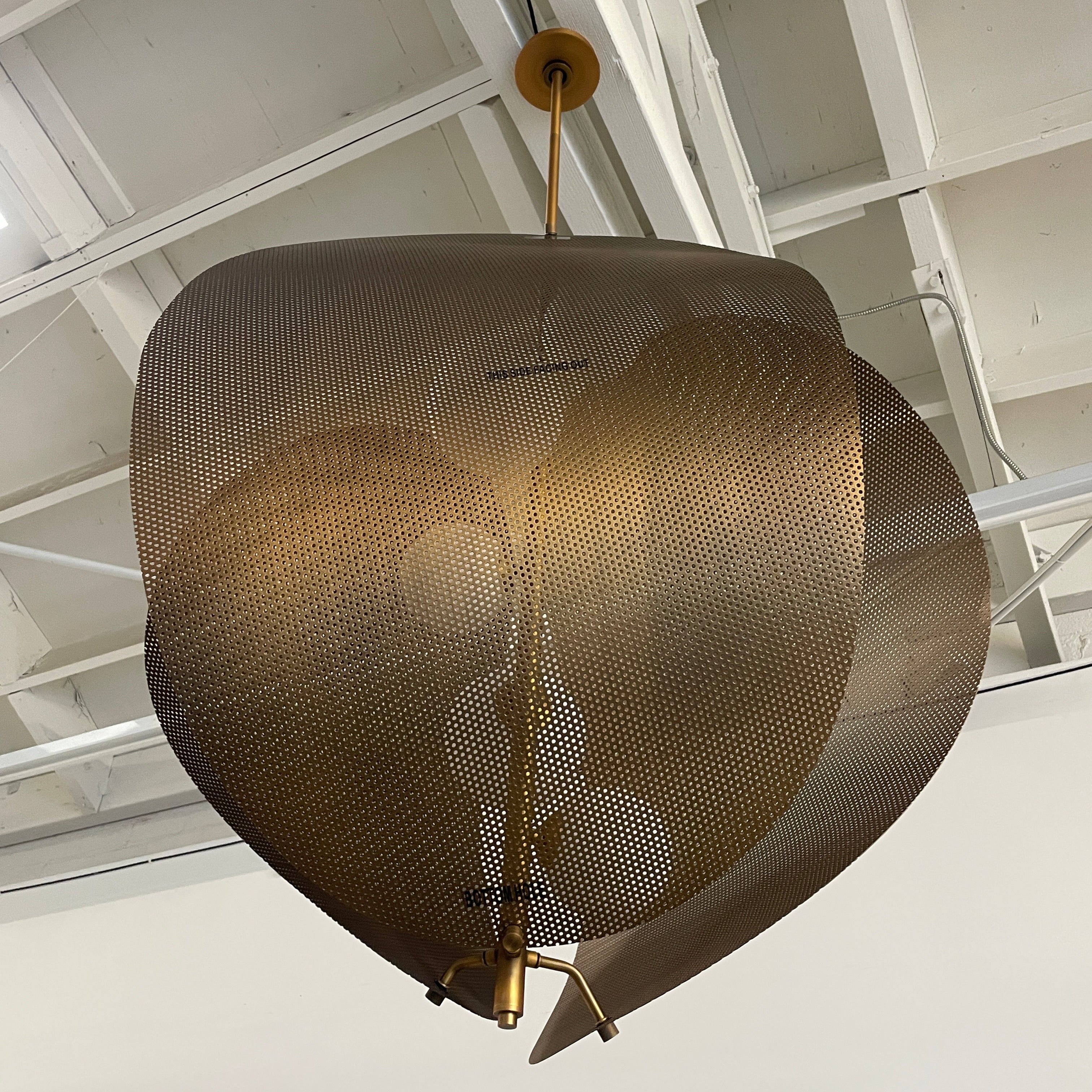 A modern chandelier with circular metal mesh panels and a central bulb hangs from a golden rod, reminiscent of the Studio M Chips 5-Light Vertical Pendant in Natural Aged Brass.