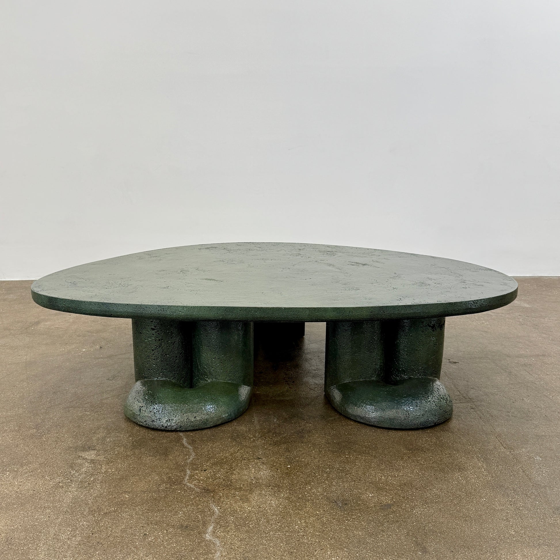 A low, irregularly shaped dark green Ian Felton Studio Mullunu Coffee Table with three thick, cylindrical legs stands on a plain white background.