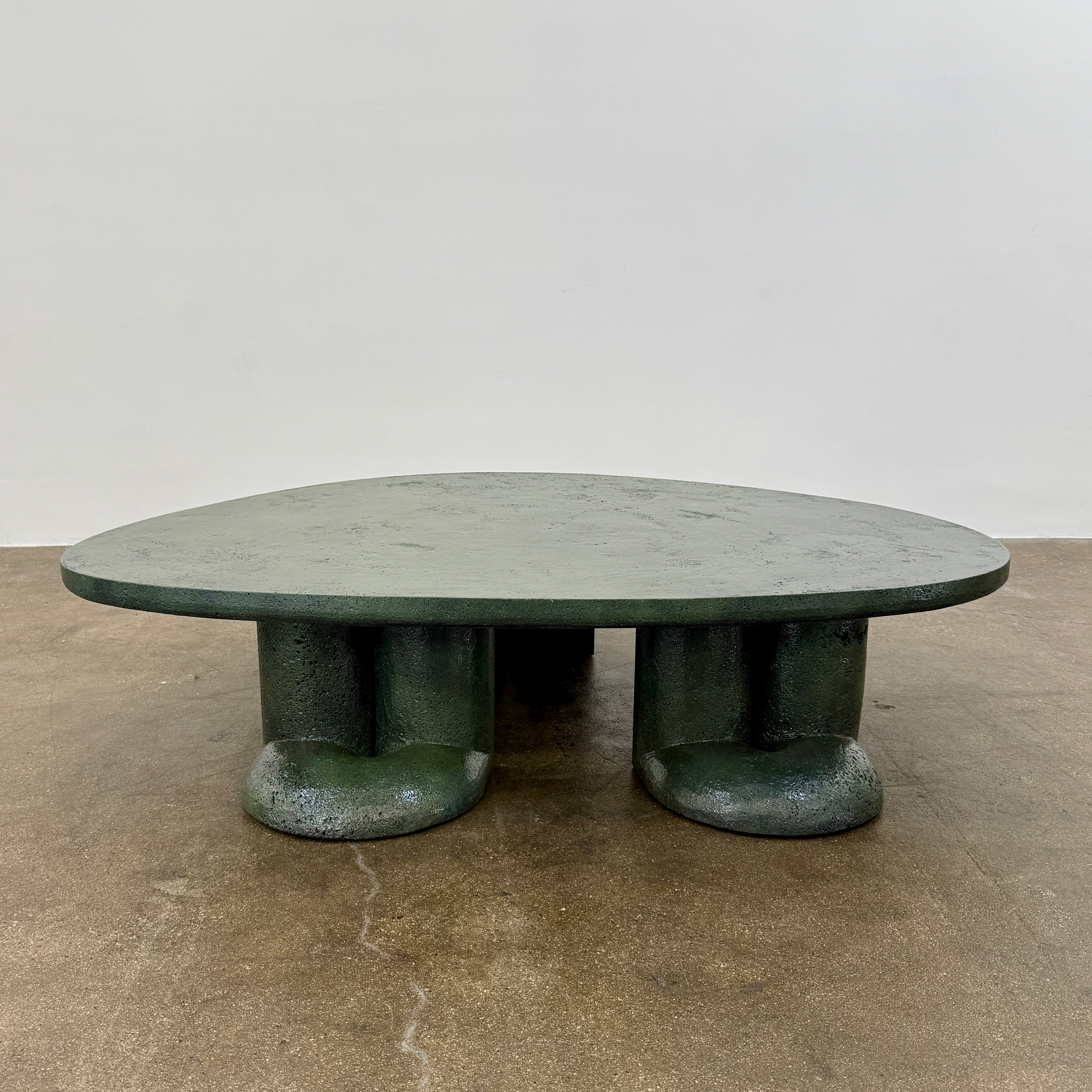 A low, irregularly shaped dark green Ian Felton Studio Mullunu Coffee Table with three thick, cylindrical legs stands on a plain white background.