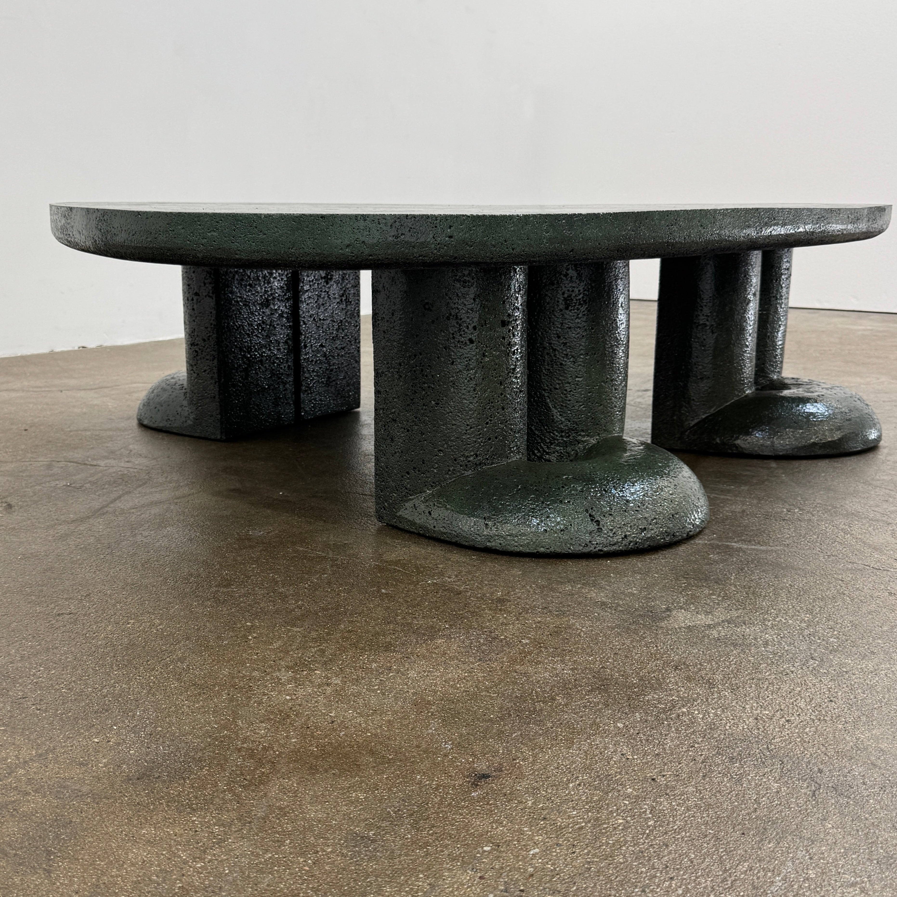 A low, irregularly shaped dark green Ian Felton Studio Mullunu Coffee Table with three thick, cylindrical legs stands on a plain white background.
