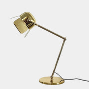 A designer table lamp with an adjustable arm stands on a white surface; a brown object clipped to the shade recalls the elegance of the Ghindini 1961 Flamingo Brass Table Lamp by Ghindini 1961.