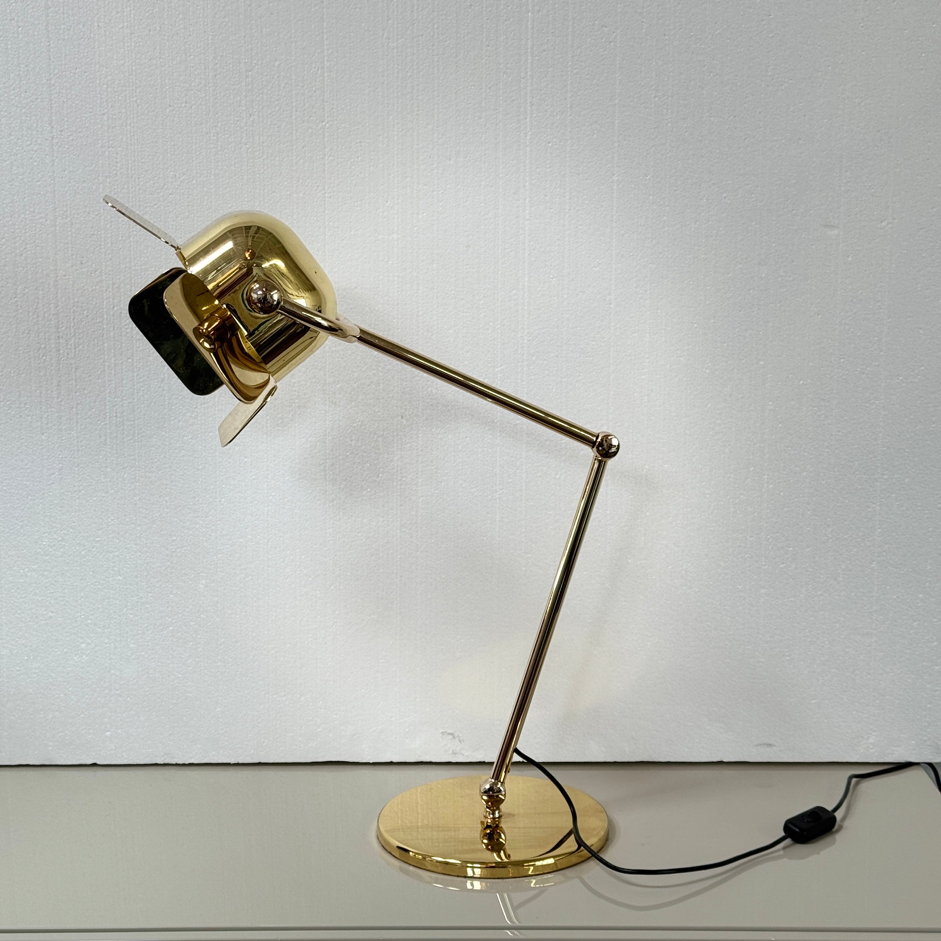 A designer table lamp with an adjustable arm stands on a white surface; a brown object clipped to the shade recalls the elegance of the Ghindini 1961 Flamingo Brass Table Lamp by Ghindini 1961.
