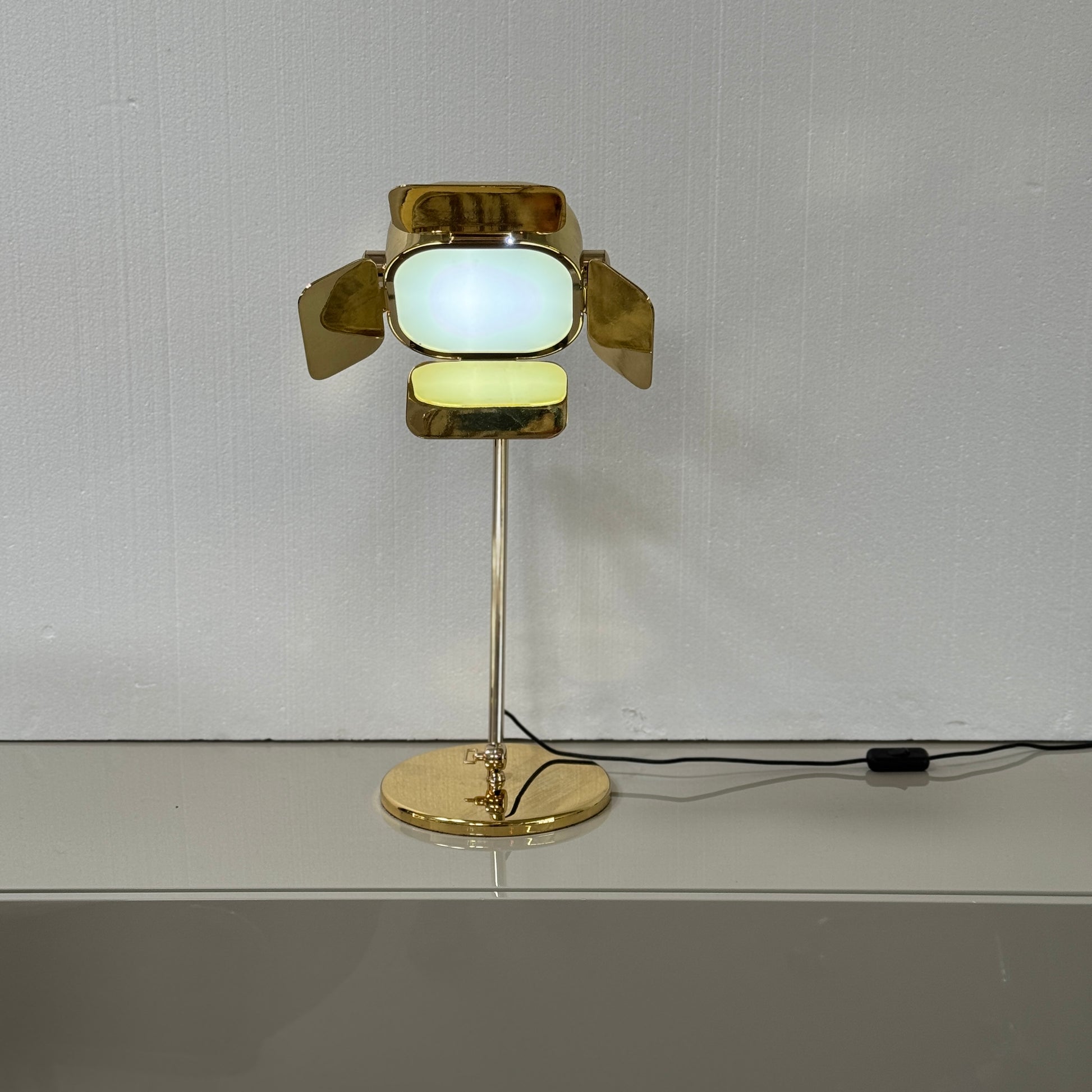 A designer table lamp with an adjustable arm stands on a white surface; a brown object clipped to the shade recalls the elegance of the Ghindini 1961 Flamingo Brass Table Lamp by Ghindini 1961.