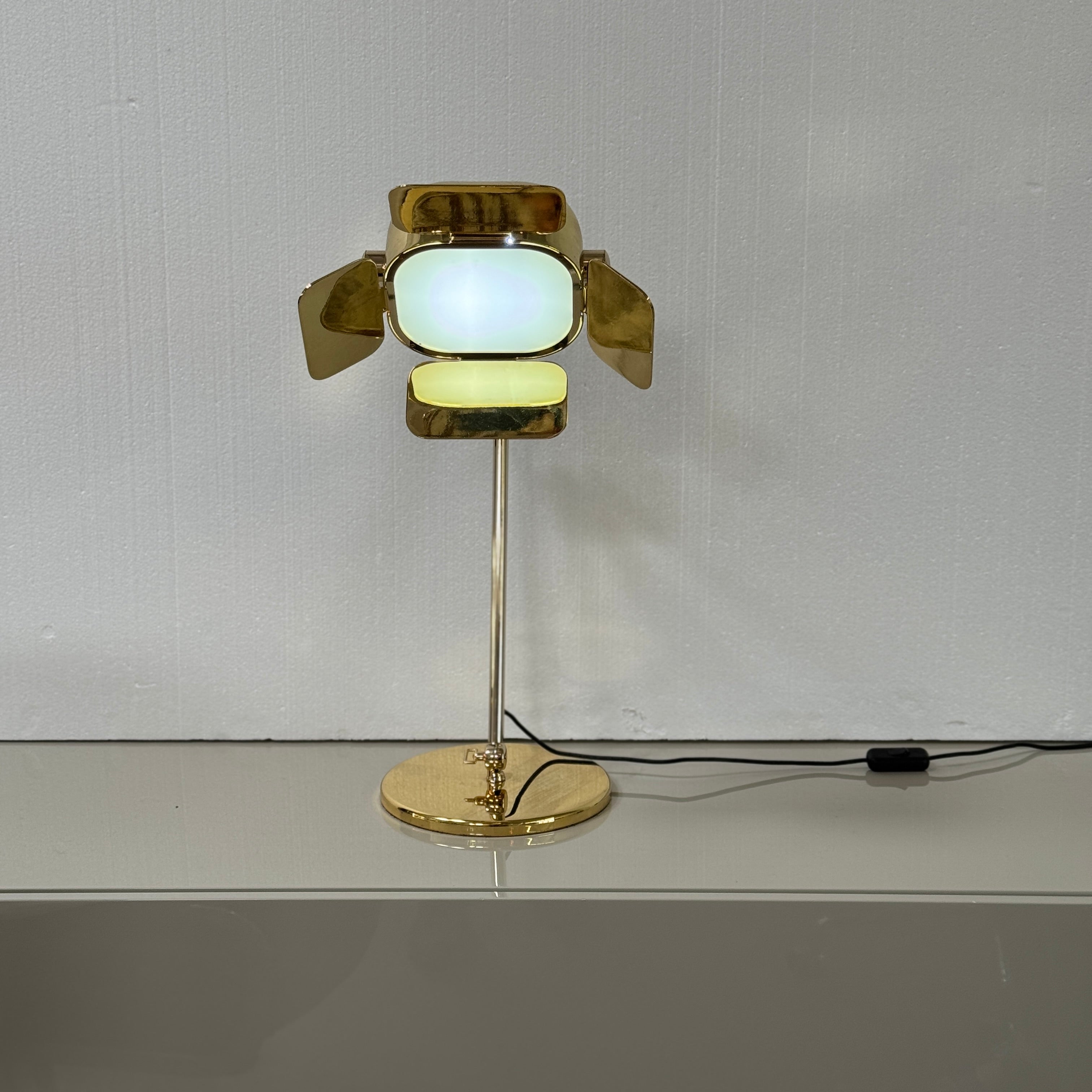 A designer table lamp with an adjustable arm stands on a white surface; a brown object clipped to the shade recalls the elegance of the Ghindini 1961 Flamingo Brass Table Lamp by Ghindini 1961.