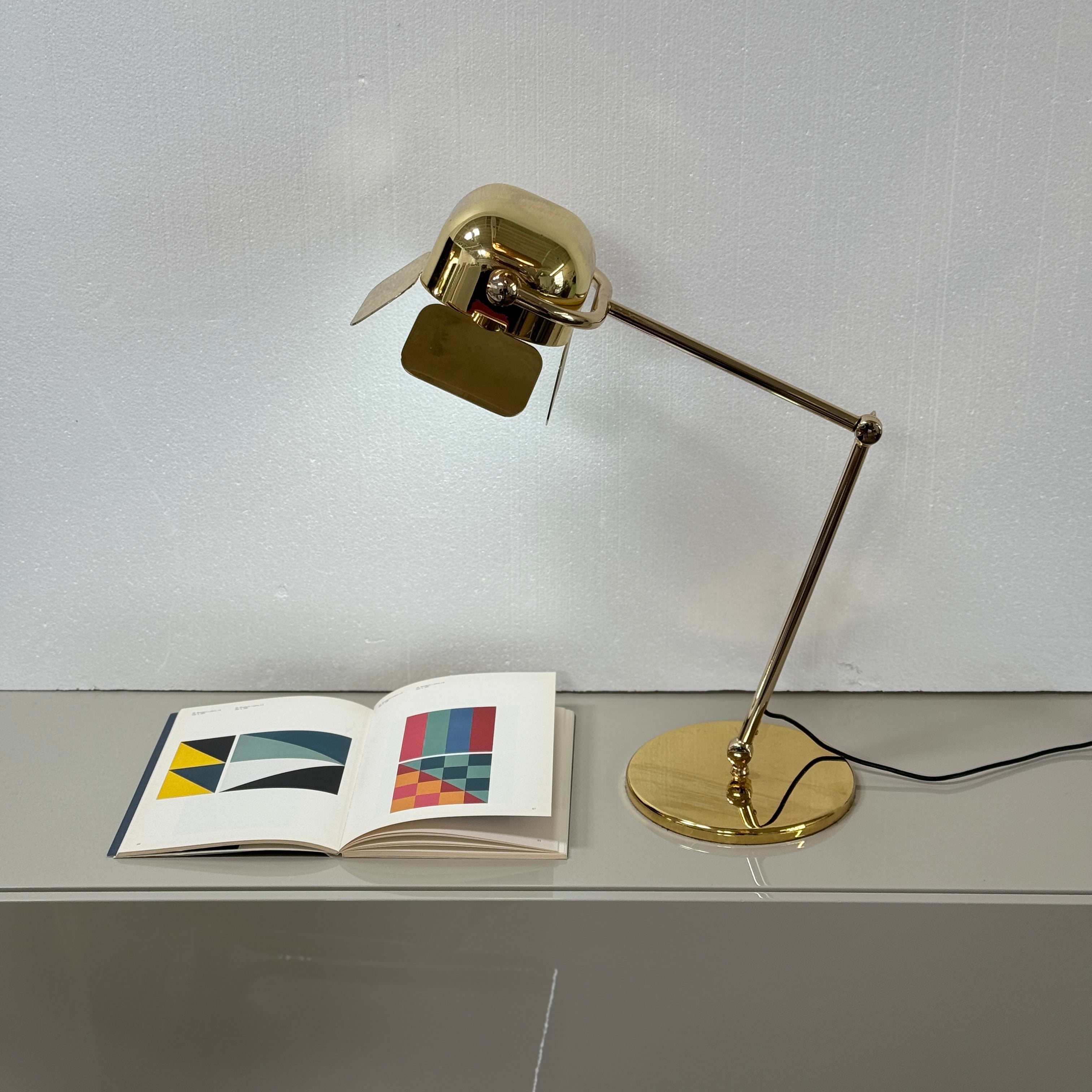 A designer table lamp with an adjustable arm stands on a white surface; a brown object clipped to the shade recalls the elegance of the Ghindini 1961 Flamingo Brass Table Lamp by Ghindini 1961.