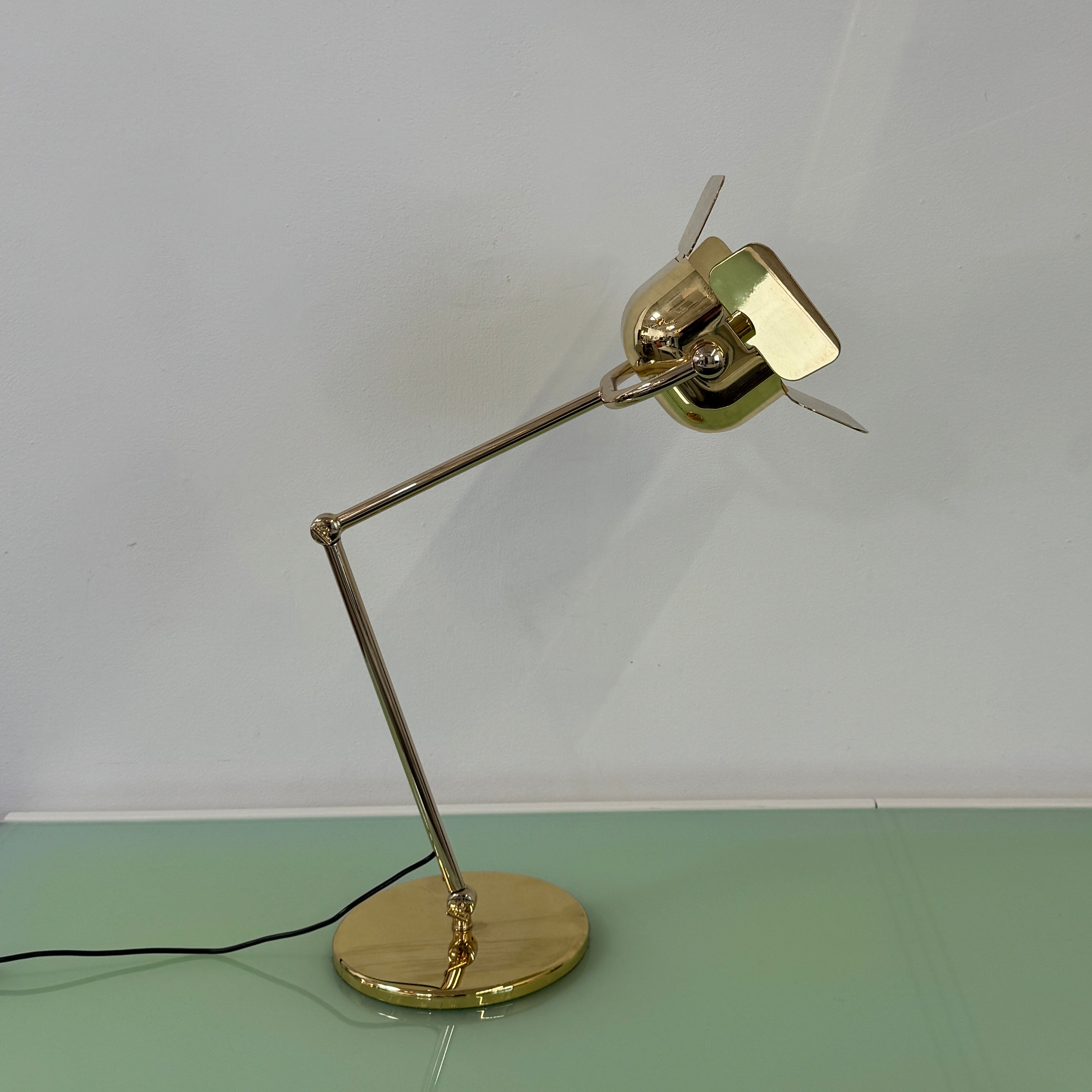 A designer table lamp with an adjustable arm stands on a white surface; a brown object clipped to the shade recalls the elegance of the Ghindini 1961 Flamingo Brass Table Lamp by Ghindini 1961.