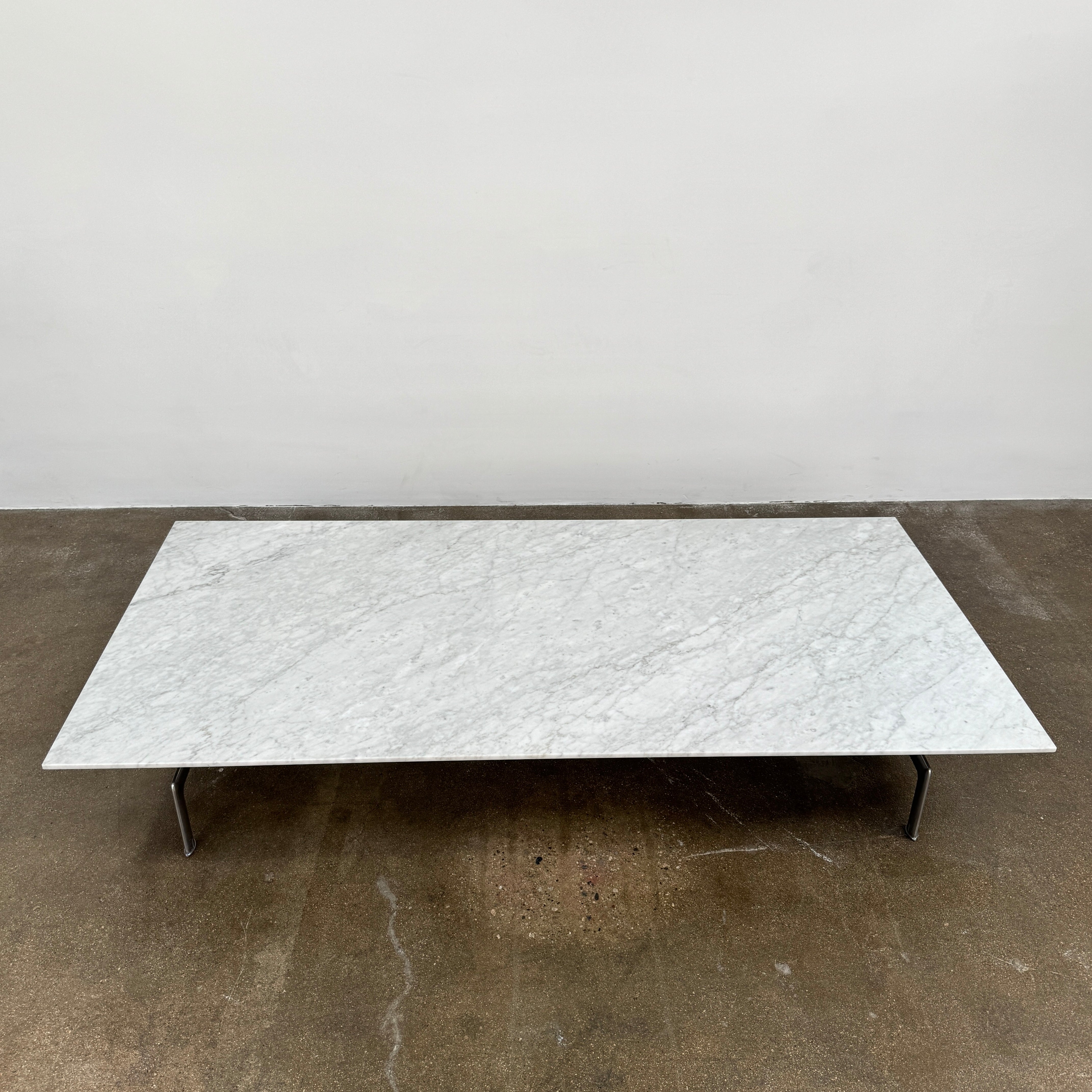 The B&B Italia Diesis Marble Coffee Table by B&B Italia, featuring a low rectangular white marble top and slim metal legs, is placed on a polished concrete floor against a plain white wall.