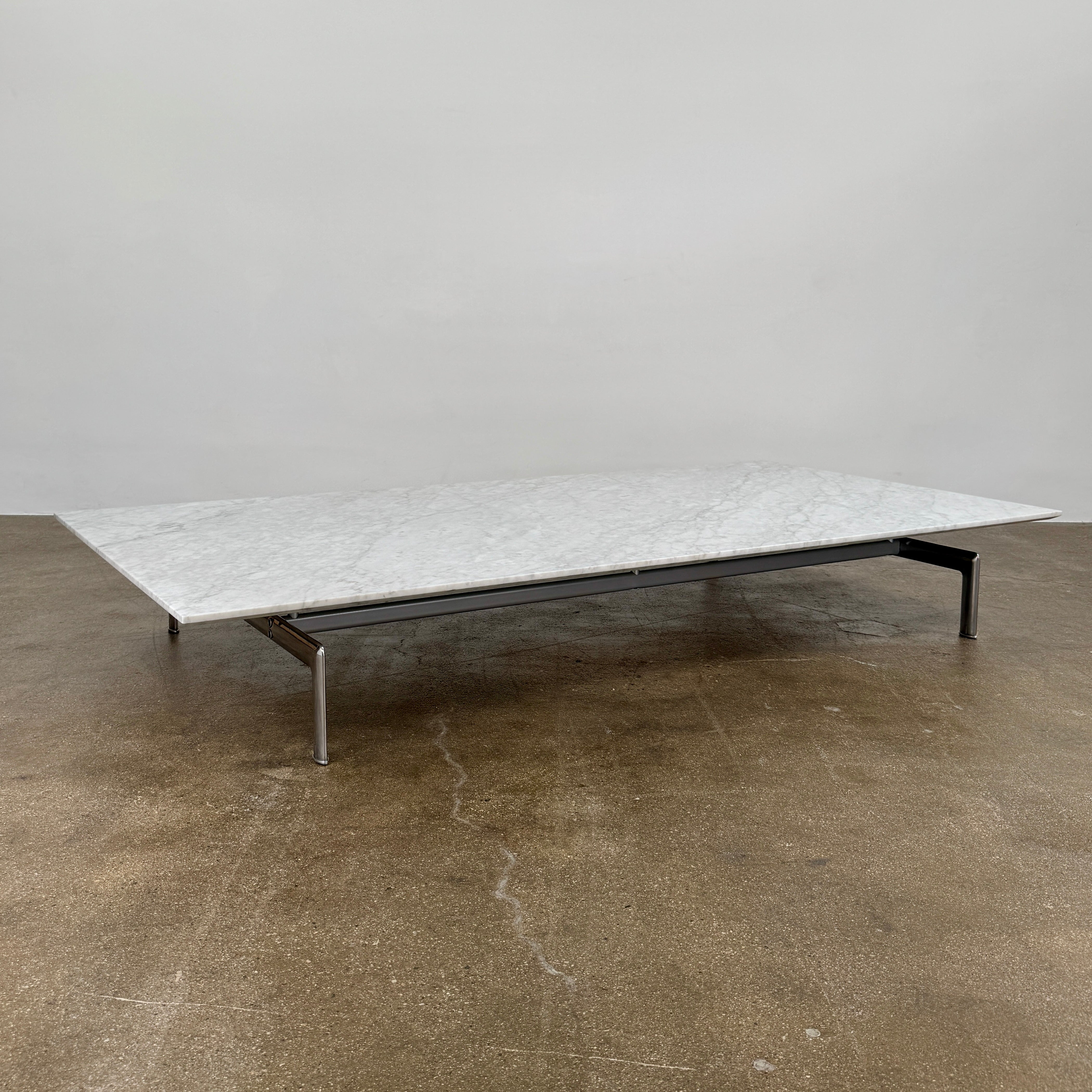 The B&B Italia Diesis Marble Coffee Table by B&B Italia, featuring a low rectangular white marble top and slim metal legs, is placed on a polished concrete floor against a plain white wall.