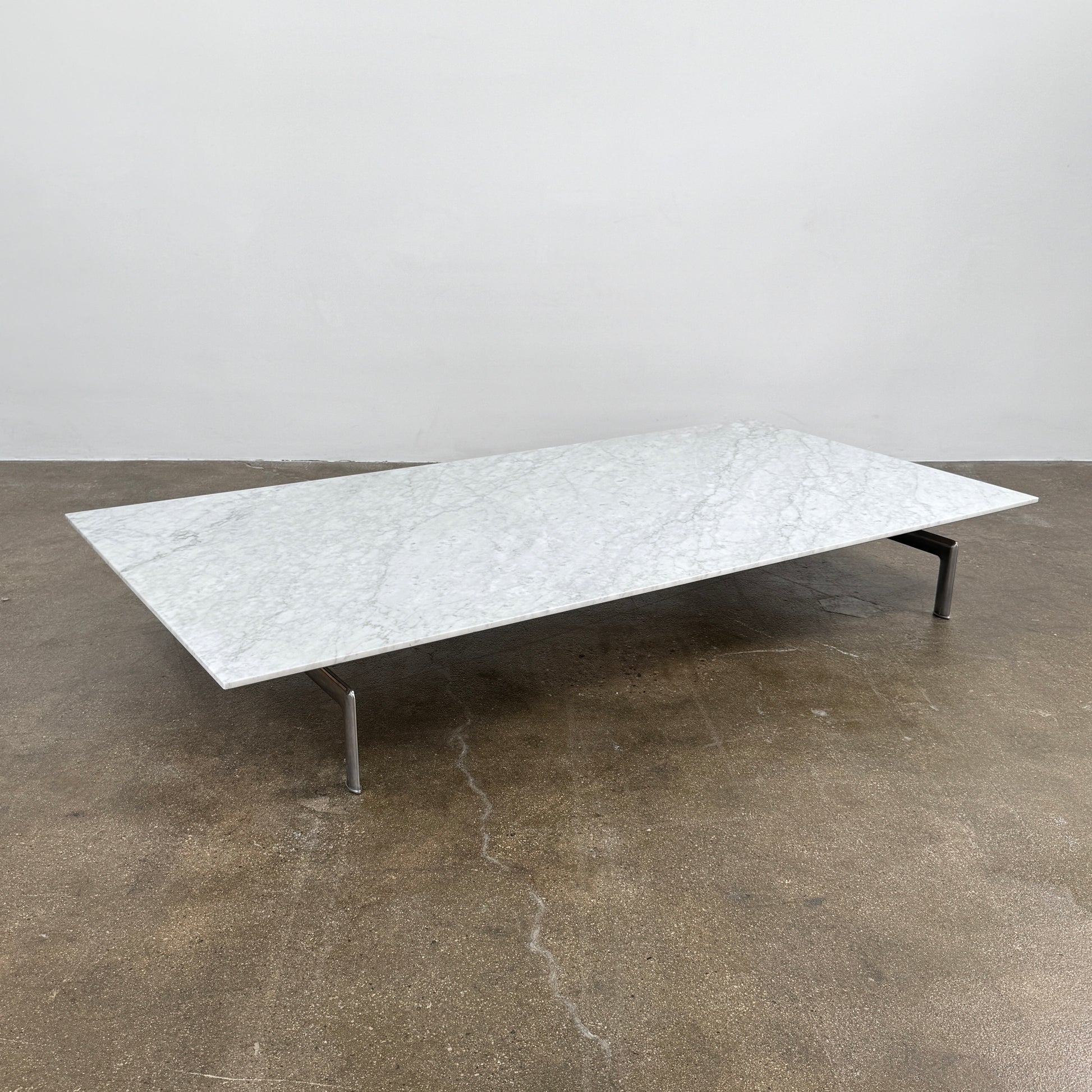 The B&B Italia Diesis Marble Coffee Table by B&B Italia, featuring a low rectangular white marble top and slim metal legs, is placed on a polished concrete floor against a plain white wall.