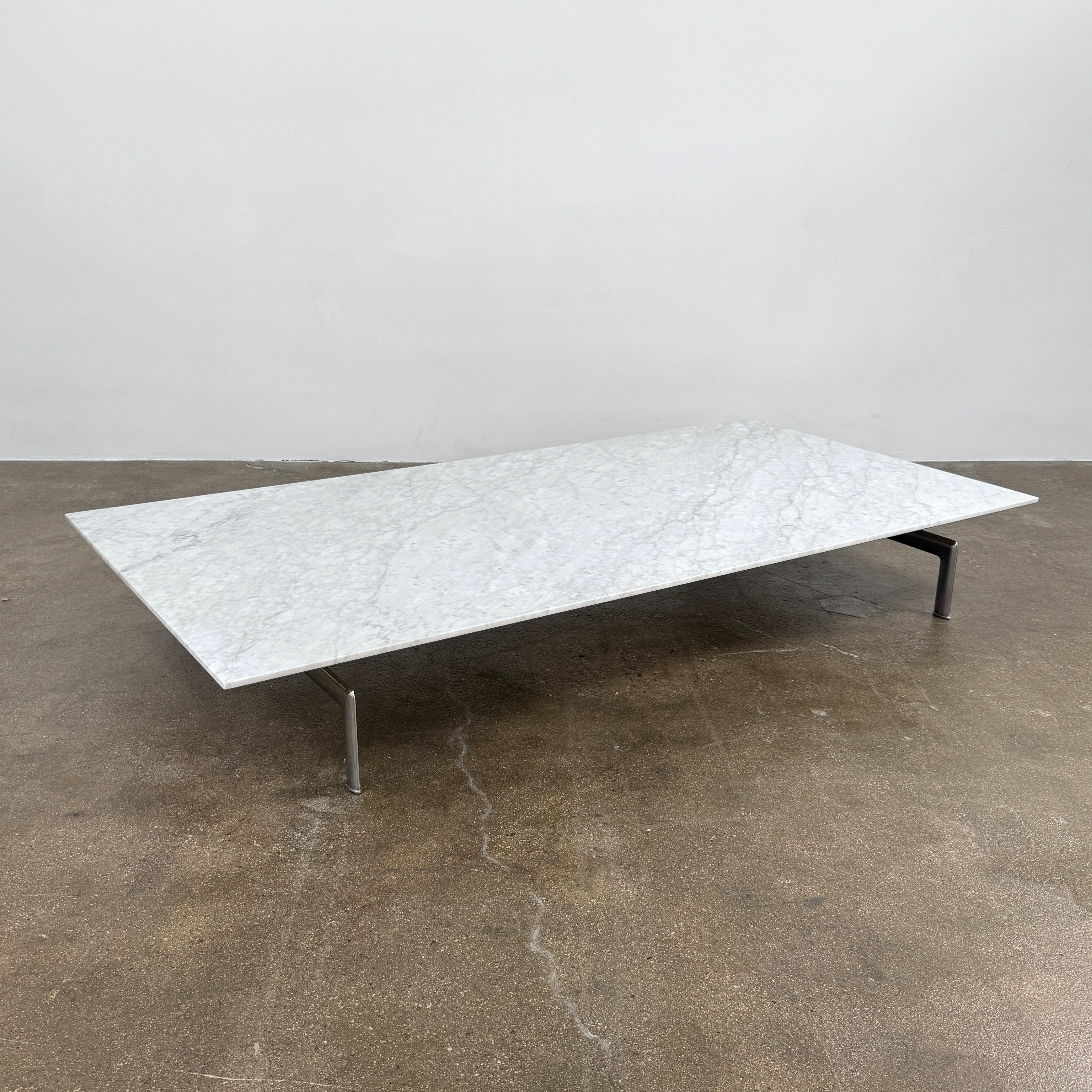 The B&B Italia Diesis Marble Coffee Table by B&B Italia, featuring a low rectangular white marble top and slim metal legs, is placed on a polished concrete floor against a plain white wall.