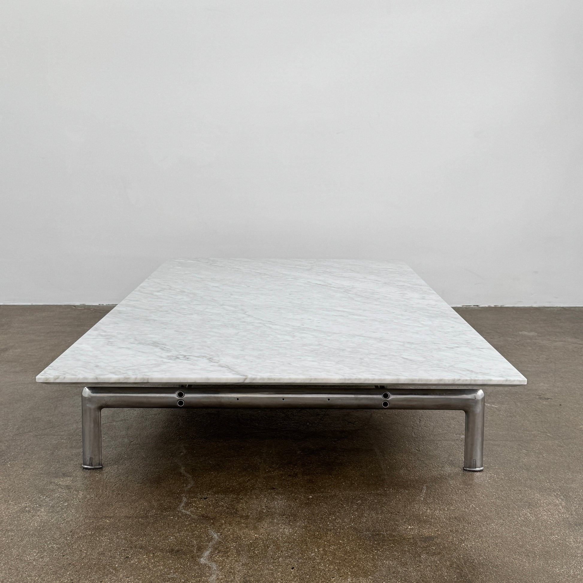 The B&B Italia Diesis Marble Coffee Table by B&B Italia, featuring a low rectangular white marble top and slim metal legs, is placed on a polished concrete floor against a plain white wall.