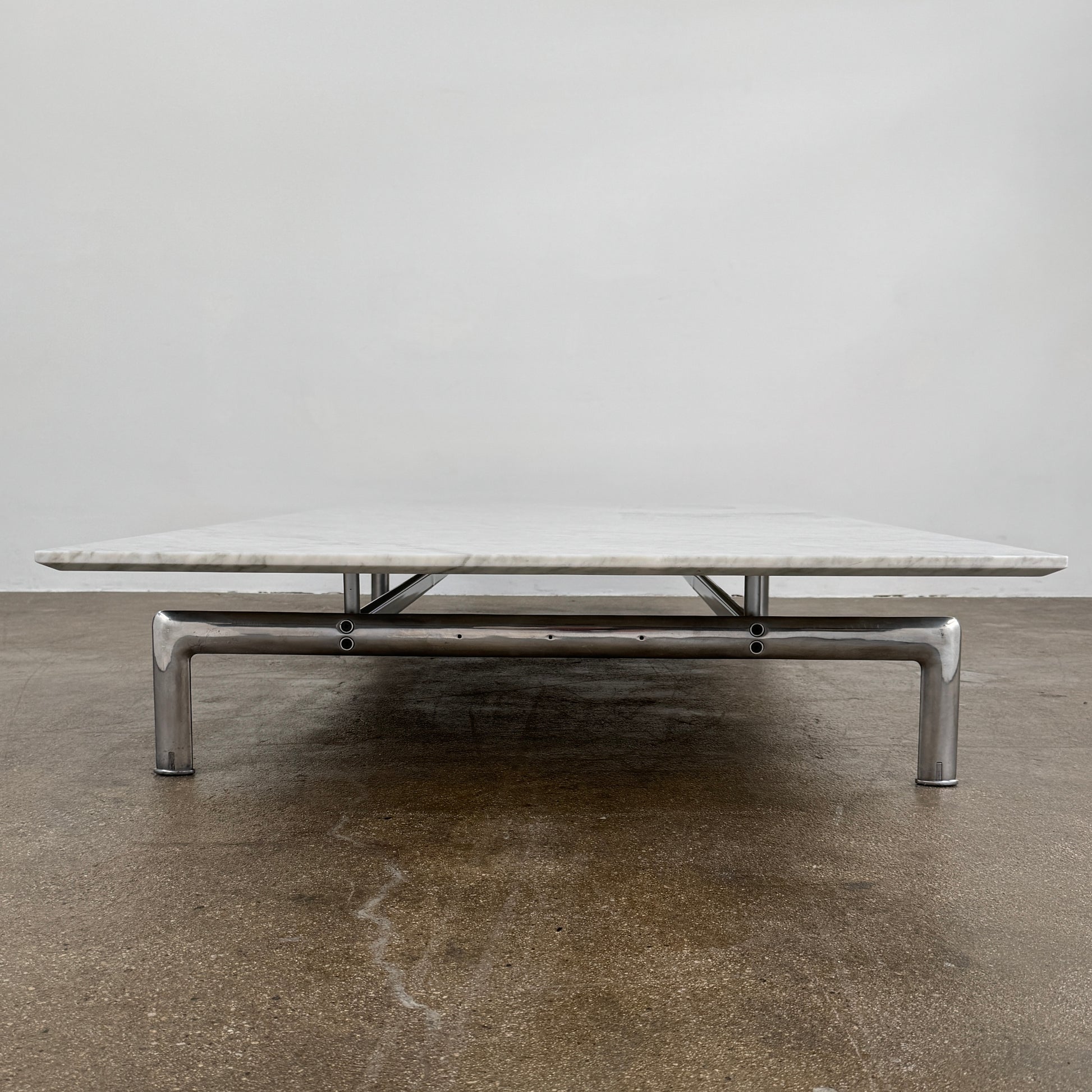 The B&B Italia Diesis Marble Coffee Table by B&B Italia, featuring a low rectangular white marble top and slim metal legs, is placed on a polished concrete floor against a plain white wall.