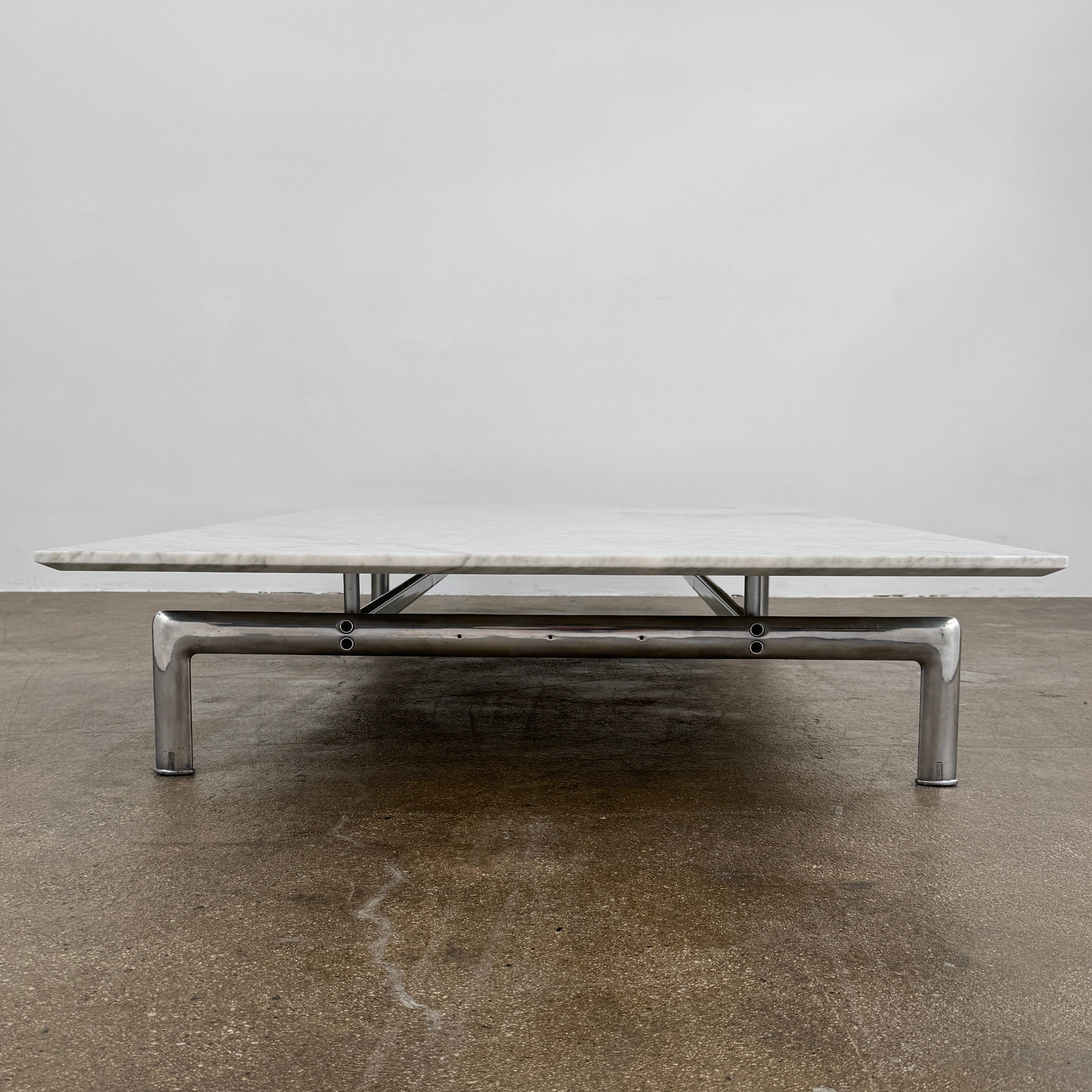 The B&B Italia Diesis Marble Coffee Table by B&B Italia, featuring a low rectangular white marble top and slim metal legs, is placed on a polished concrete floor against a plain white wall.