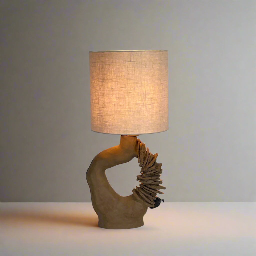 The Sofi Ceramic Arch Lamp by Sofi Ceramics is a distinctively designed table lamp with a twisting sculptural base and a cylindrical shade. Its hand-built ceramic elements softly illuminate against plain backgrounds.