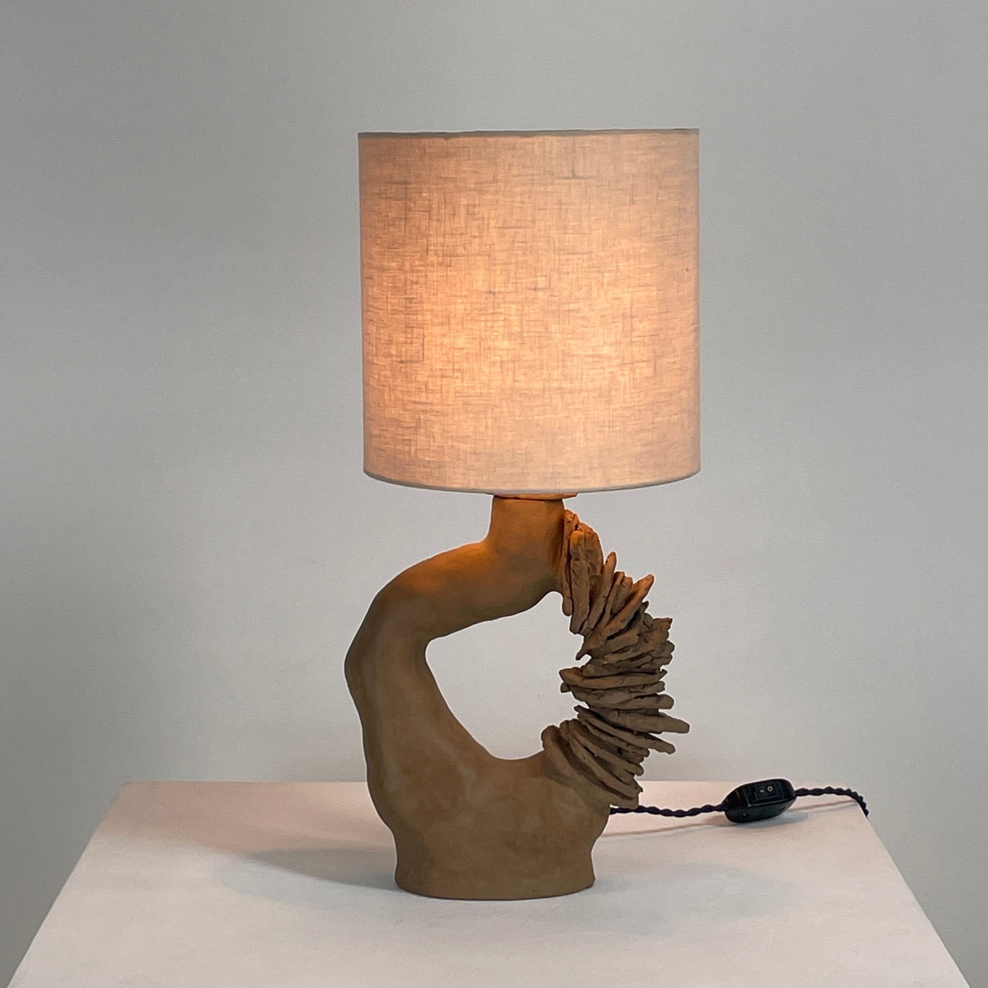 The Sofi Ceramic Arch Lamp by Sofi Ceramics is a distinctively designed table lamp with a twisting sculptural base and a cylindrical shade. Its hand-built ceramic elements softly illuminate against plain backgrounds.