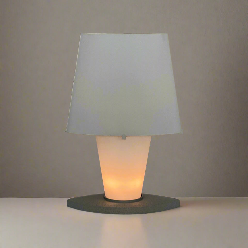 The Fontana Arte 3064 Table Light by Fontana Arte, echoing Daniela Puppa's designs, boasts a wide conical shade for a warm glow. It sits elegantly on a white surface against a plain backdrop, capturing the essence of postmodern art.