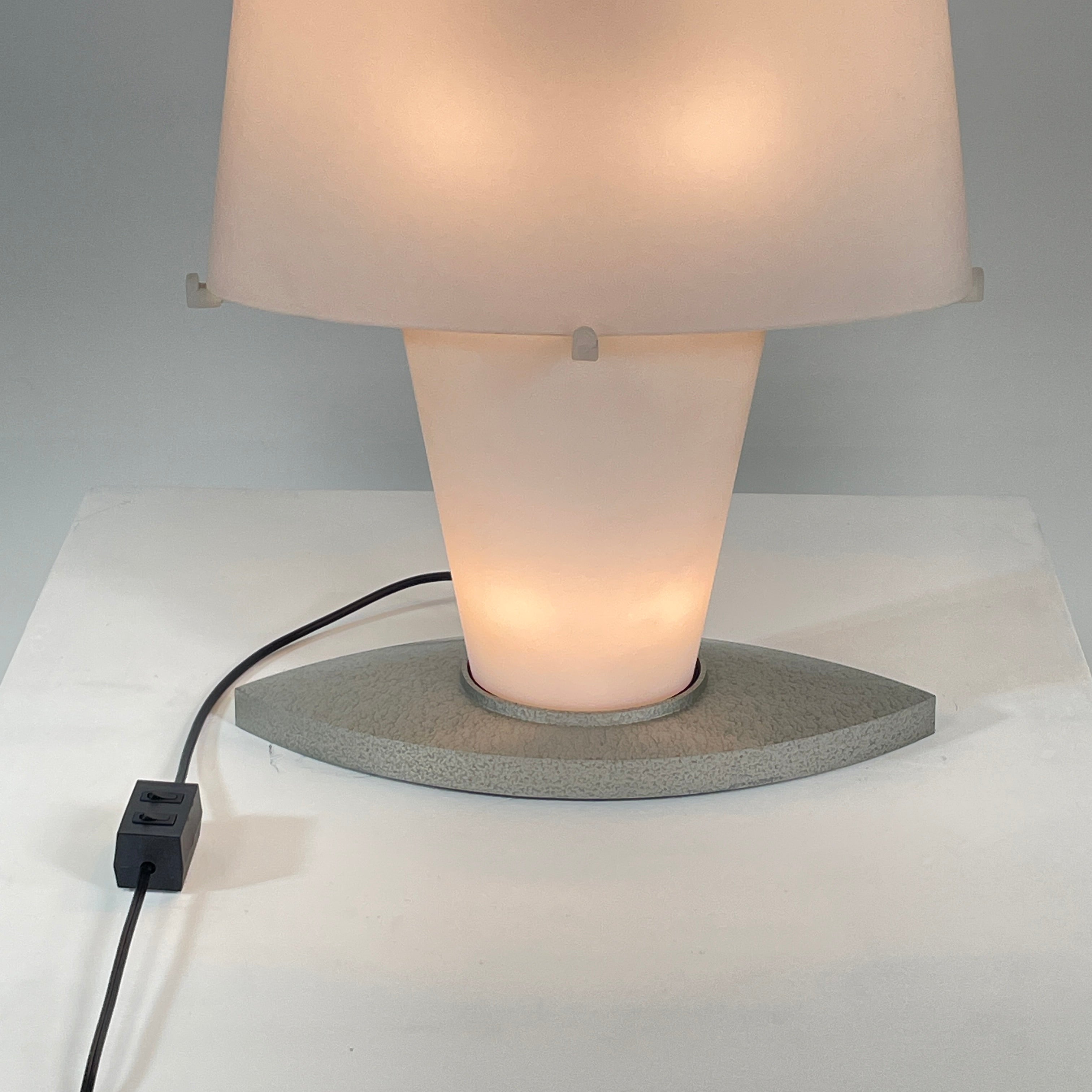 The Fontana Arte 3064 Table Light by Fontana Arte, echoing Daniela Puppa's designs, boasts a wide conical shade for a warm glow. It sits elegantly on a white surface against a plain backdrop, capturing the essence of postmodern art.