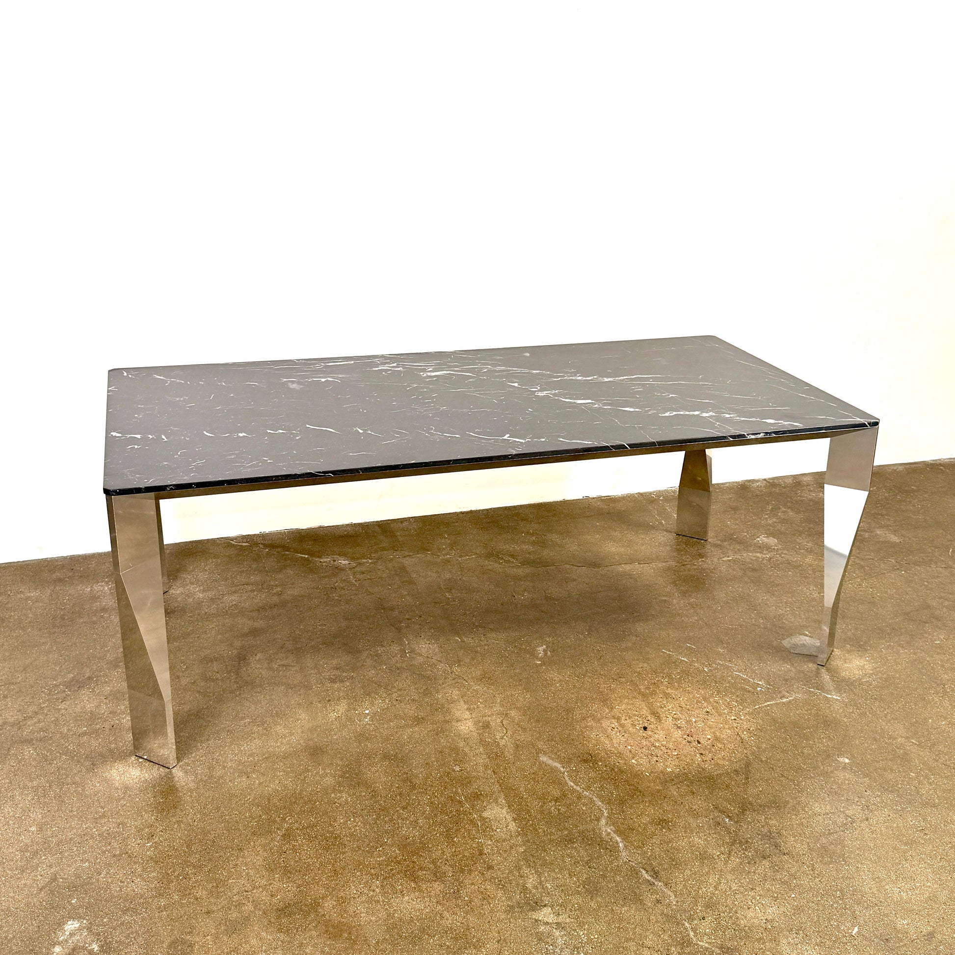 The Molteni & C Diamond table features a rectangular black marble top and angled polished metal legs, set on a brown concrete floor against a plain white wall. This piece by Molteni & C adds sophisticated elegance to any space.
