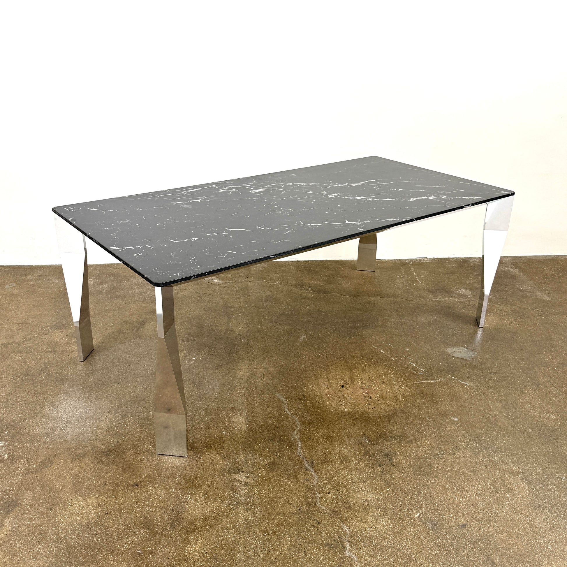 The Molteni & C Diamond table features a rectangular black marble top and angled polished metal legs, set on a brown concrete floor against a plain white wall. This piece by Molteni & C adds sophisticated elegance to any space.