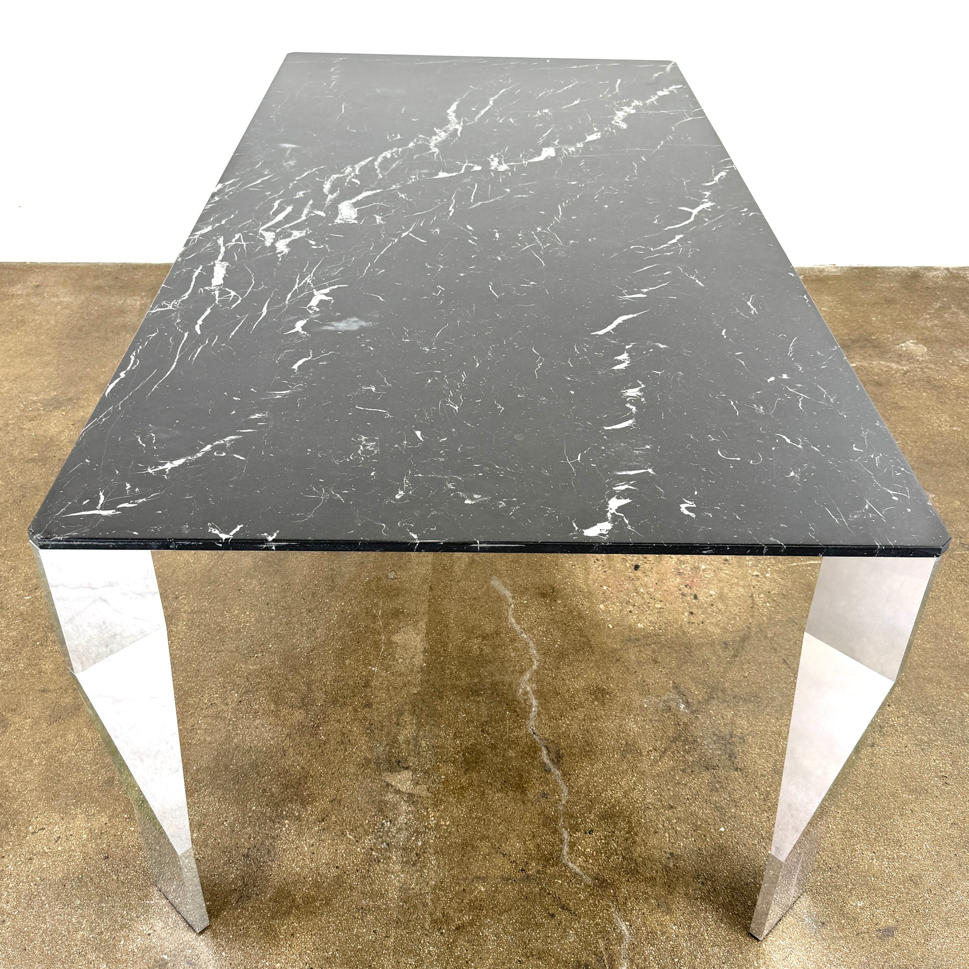 The Molteni & C Diamond table features a rectangular black marble top and angled polished metal legs, set on a brown concrete floor against a plain white wall. This piece by Molteni & C adds sophisticated elegance to any space.