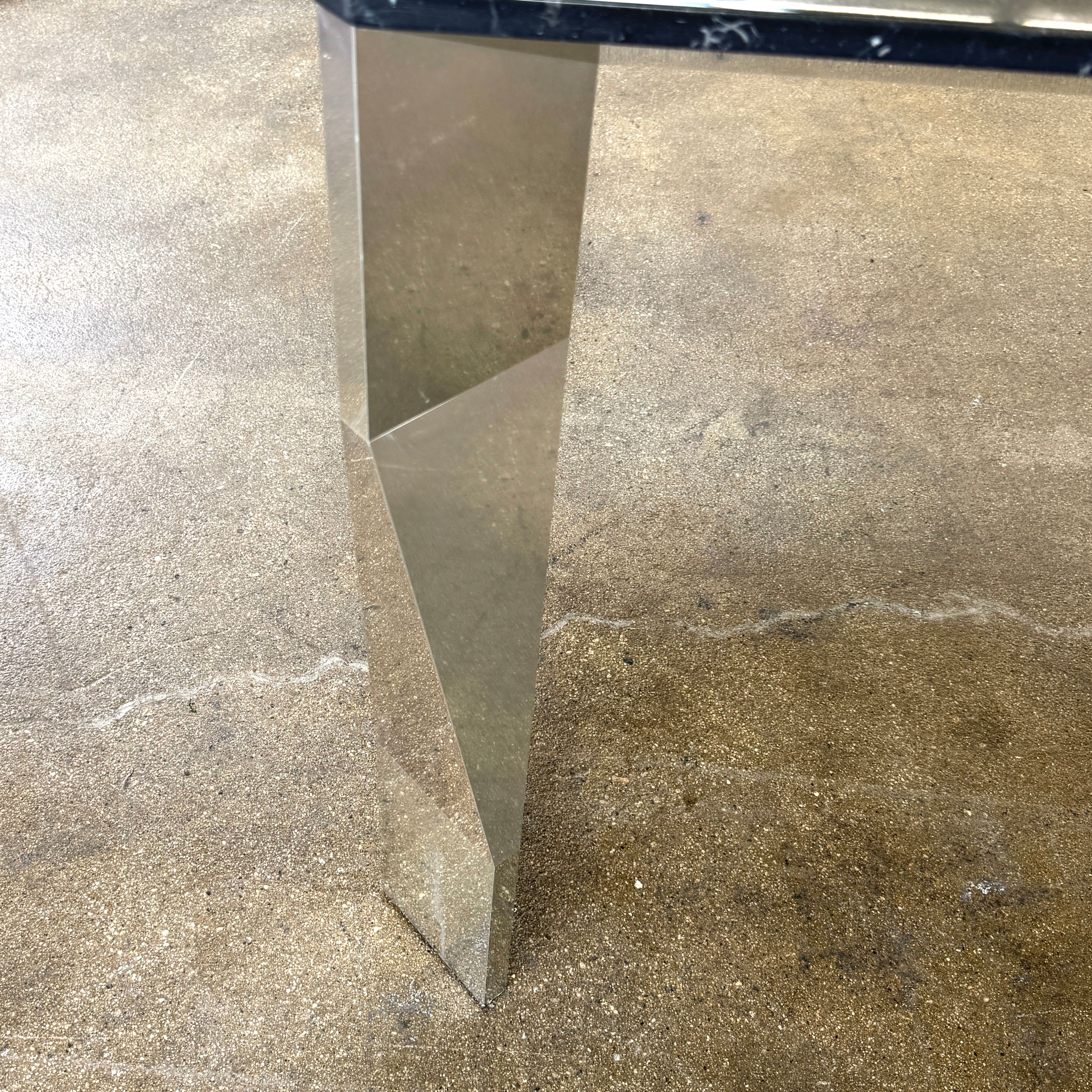 The Molteni & C Diamond table features a rectangular black marble top and angled polished metal legs, set on a brown concrete floor against a plain white wall. This piece by Molteni & C adds sophisticated elegance to any space.