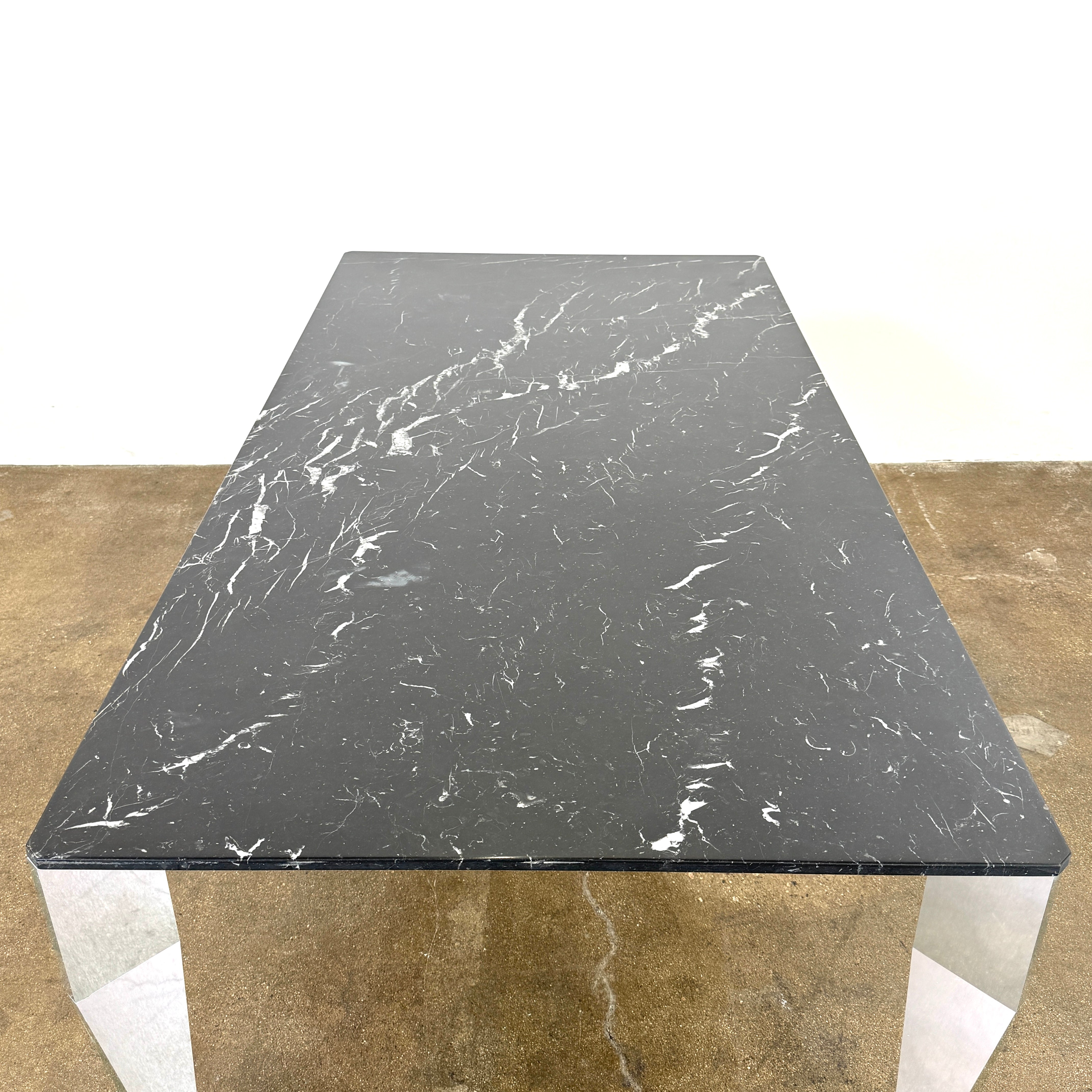 The Molteni & C Diamond table features a rectangular black marble top and angled polished metal legs, set on a brown concrete floor against a plain white wall. This piece by Molteni & C adds sophisticated elegance to any space.