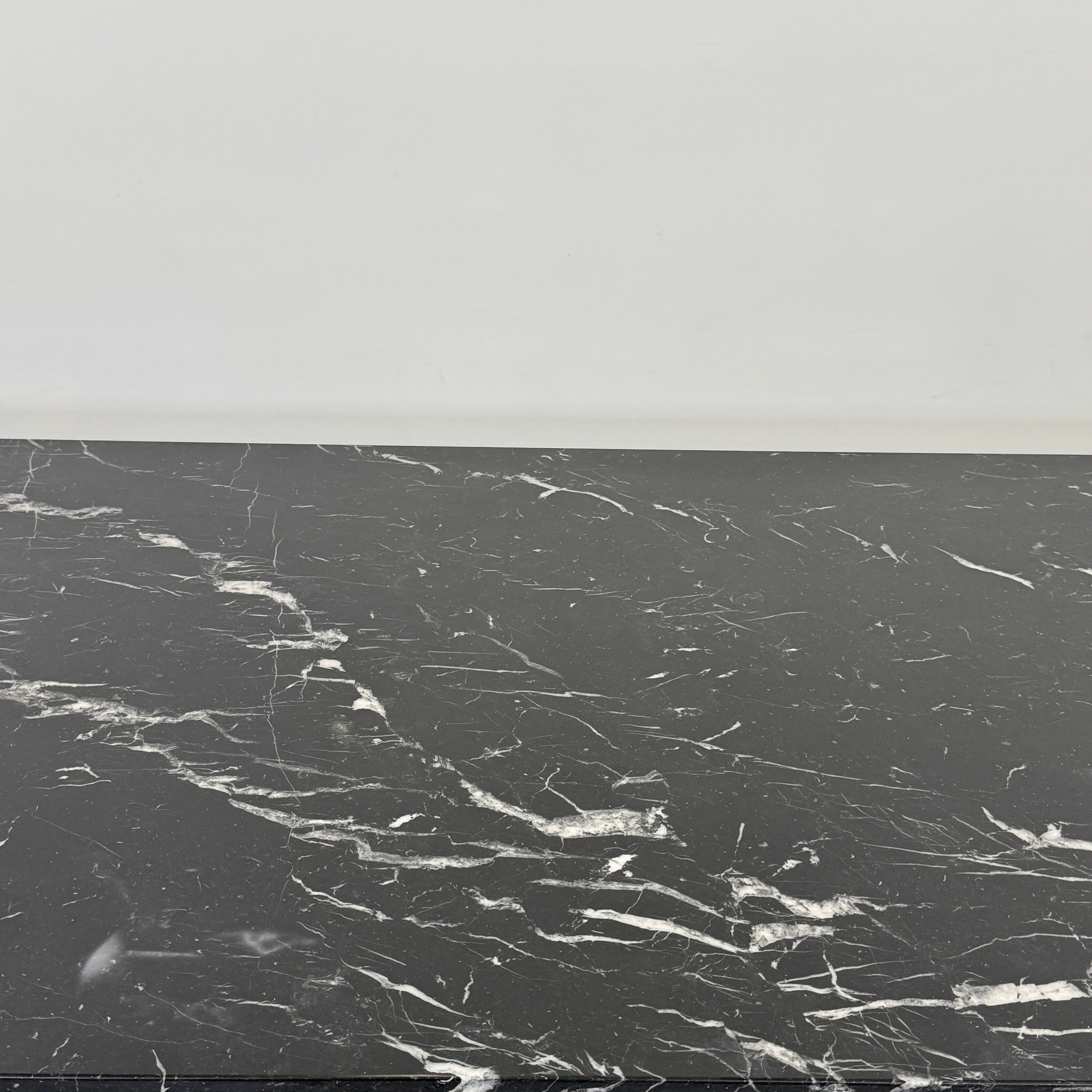 The Molteni & C Diamond table features a rectangular black marble top and angled polished metal legs, set on a brown concrete floor against a plain white wall. This piece by Molteni & C adds sophisticated elegance to any space.