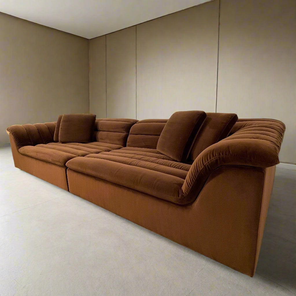 The Ellison Studios Float Sofa is a dark brown sectional from Ellison Studios with velvet upholstery, low profile, channel tufting, and two large cushions for extra comfort.