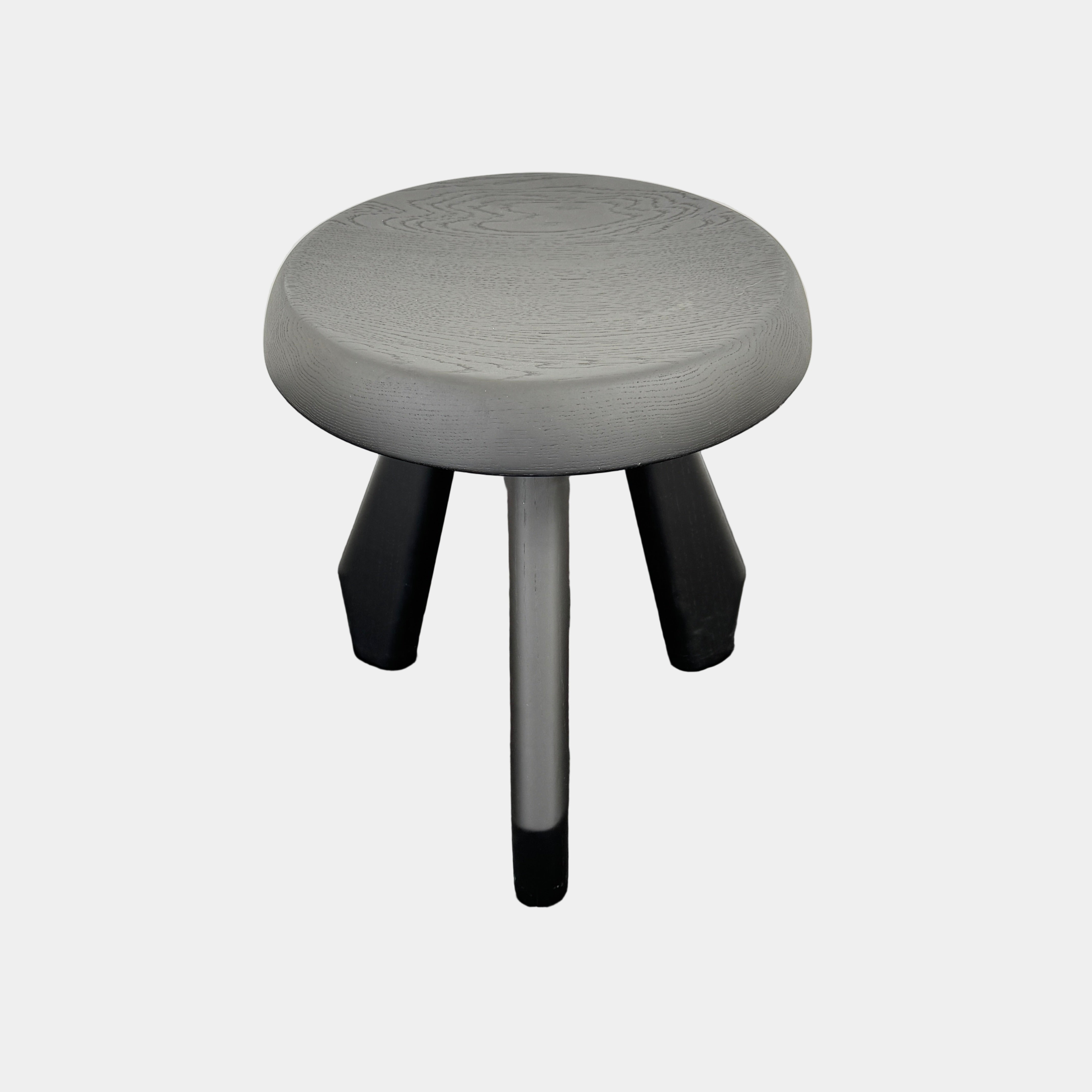 The Cassina 523 Tabouret Meribel by Cassina is a small, round, three-legged solid wood stool with a gray seat and black legs, shown against a plain white background.