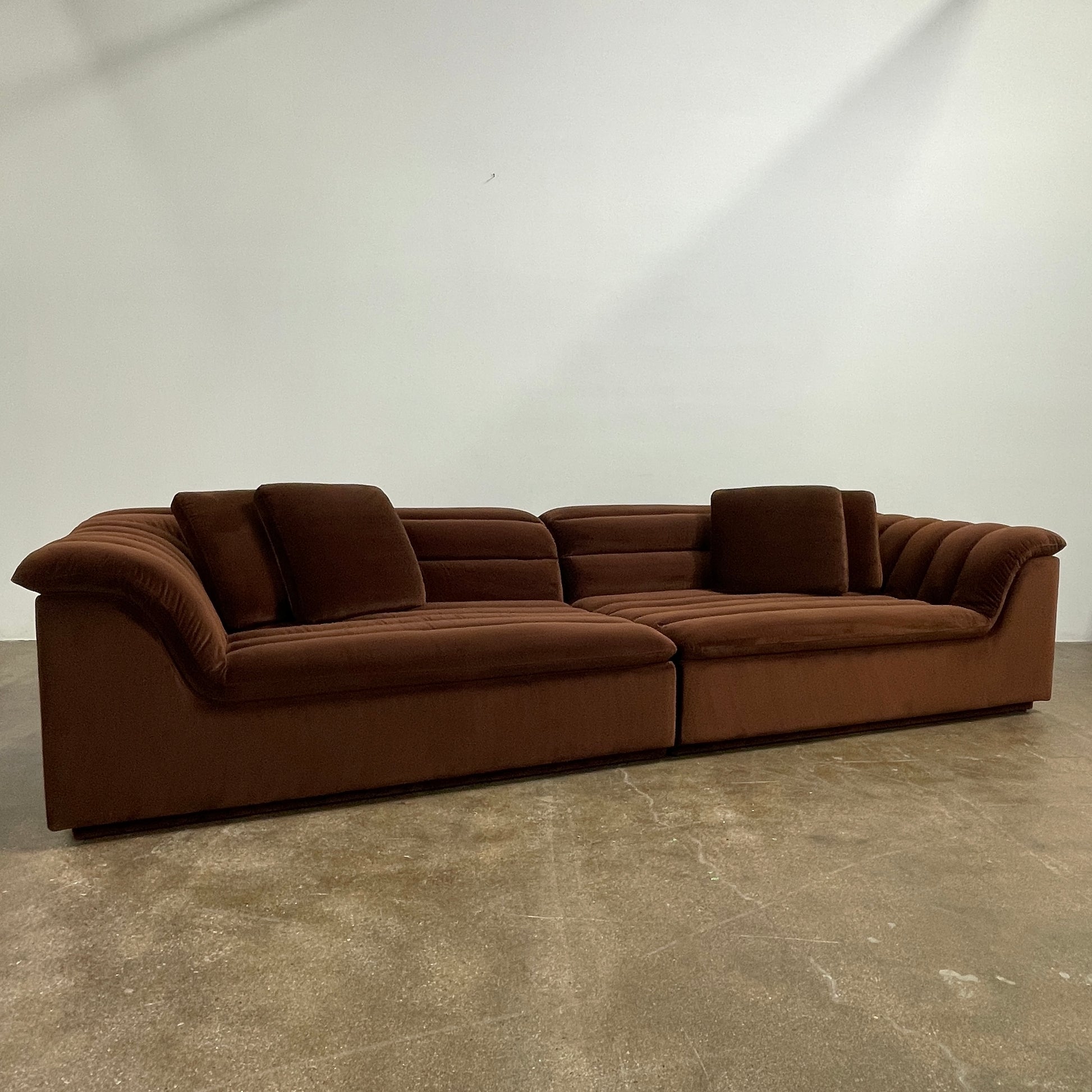 The Ellison Studios Float Sofa is a dark brown sectional from Ellison Studios with velvet upholstery, low profile, channel tufting, and two large cushions for extra comfort.
