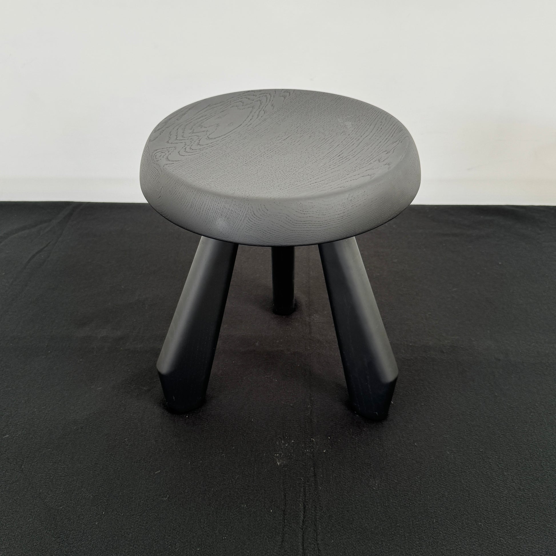 The Cassina 523 Tabouret Meribel by Cassina is a small, round, three-legged solid wood stool with a gray seat and black legs, shown against a plain white background.