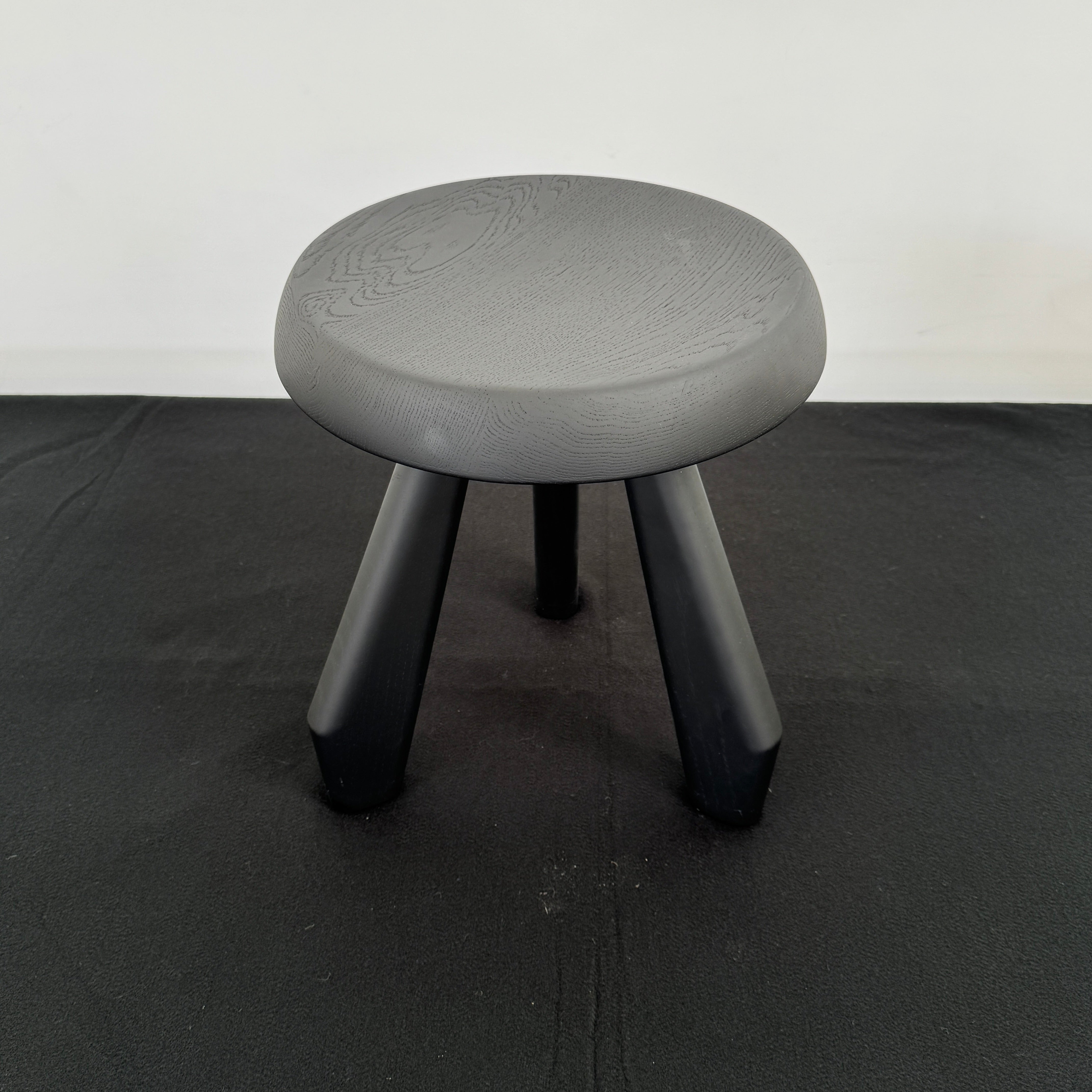 The Cassina 523 Tabouret Meribel by Cassina is a small, round, three-legged solid wood stool with a gray seat and black legs, shown against a plain white background.